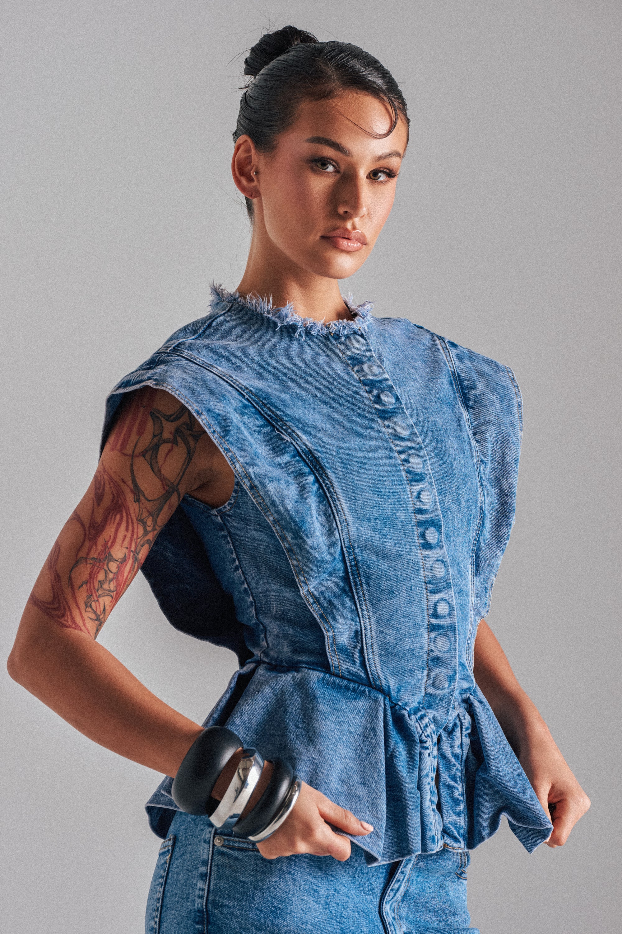 A tattooed woman with her hair in a bun poses confidently in the DONT PLAY WITH MY HEART DENIM BLOUSE, featuring distressed details and frayed edges, styled with blue jeans and bold black and silver bangles.