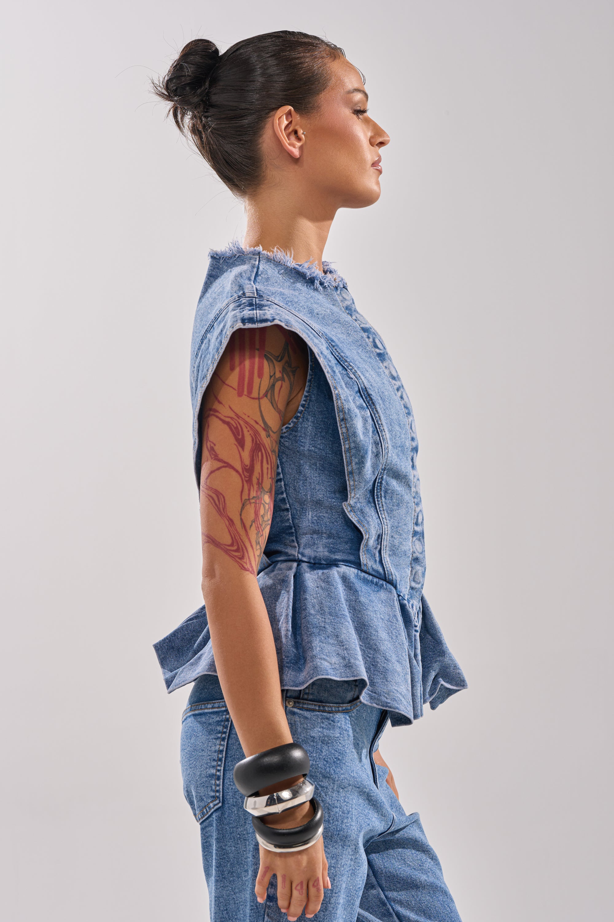 A woman with a bun, arm tattoo, and chunky bracelets poses in profile wearing the DONT PLAY WITH MY HEART DENIM BLOUSE and matching jeans against a neutral background.