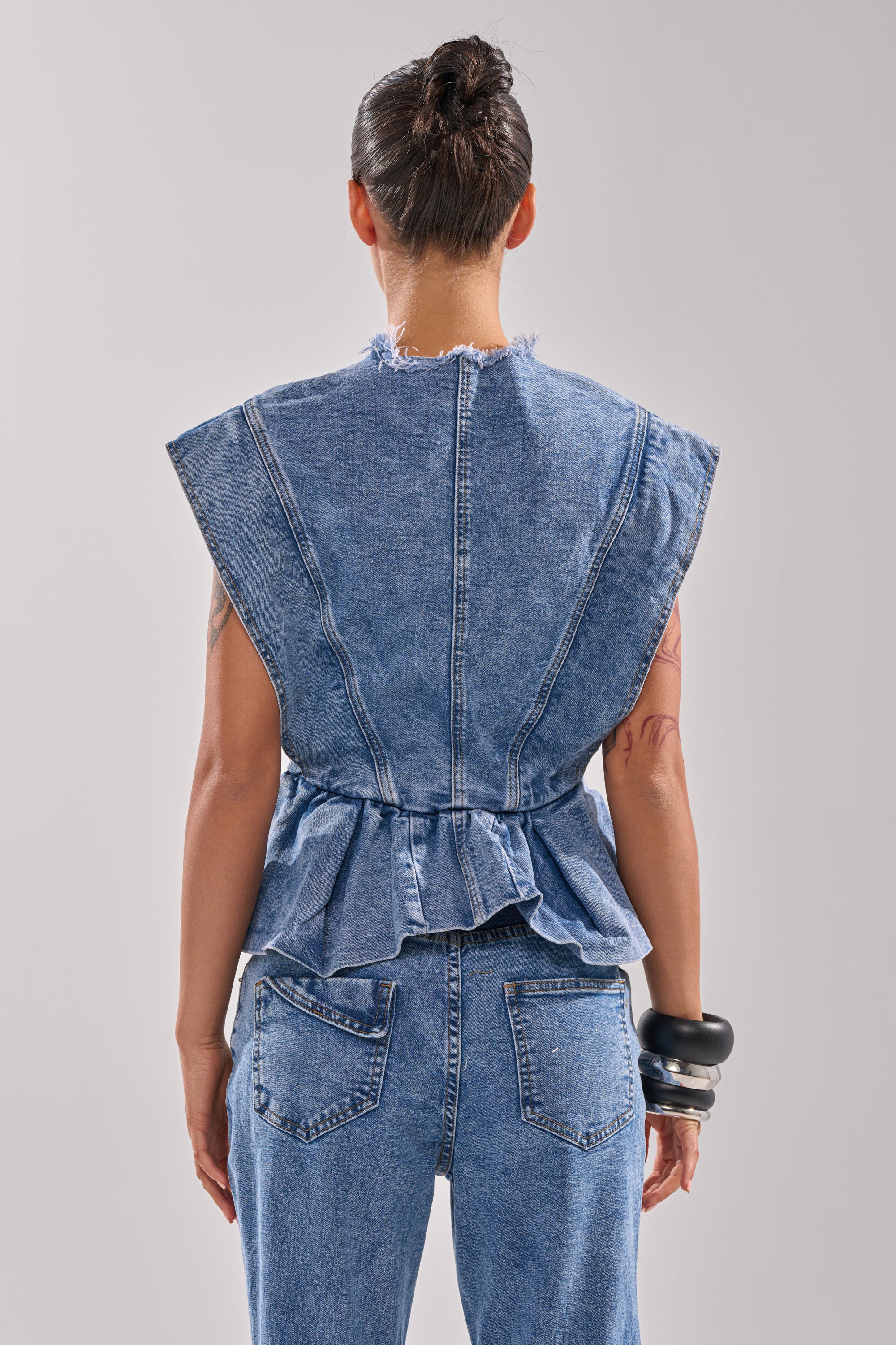 A person with dark hair in a bun wears the DONT PLAY WITH MY HEART DENIM BLOUSE, a sleeveless belted top with a ruffled peplum hem, paired with matching jeans. Tattoos cover their arms and chunky bracelets adorn their right wrist. Shown from the back.
