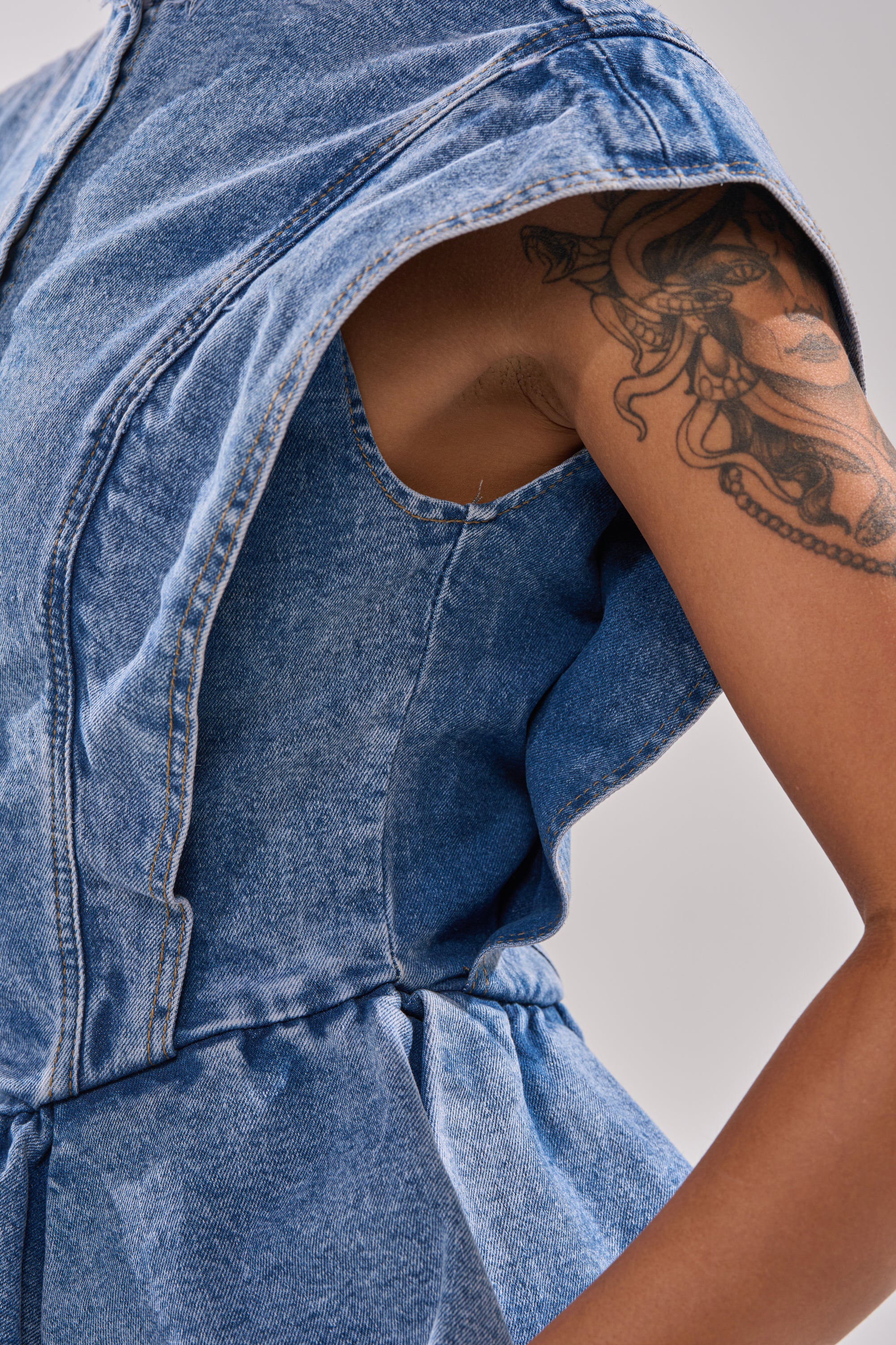 A person wears the DONT PLAY WITH MY HEART DENIM BLOUSE—a sleeveless blue denim top with distressed details—showing a tattooed upper arm, against a plain light background.