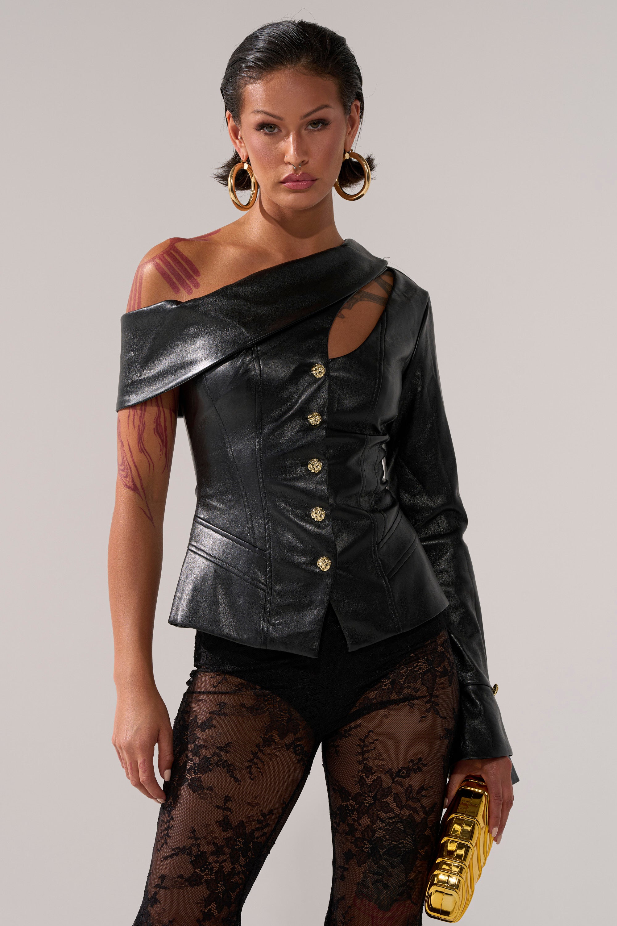 A person wears the YOU GOT ME FEELING FAUX LEATHER BUTTON DOWN with gold buttons, sheer black lace pants, large hoop earrings, and a gold clutch against a plain backdrop.