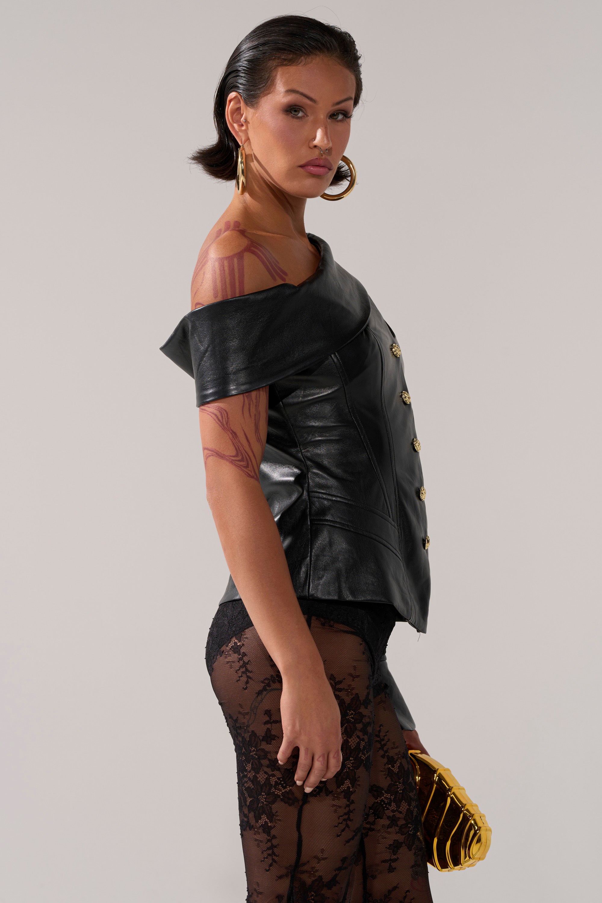 A woman with slicked-back dark hair wears the YOU GOT ME FEELING FAUX LEATHER BUTTON DOWN off-shoulder top, black lace pants, gold hoop earrings, and holds a gold clutch. She has arm tattoos and looks confidently at the camera.