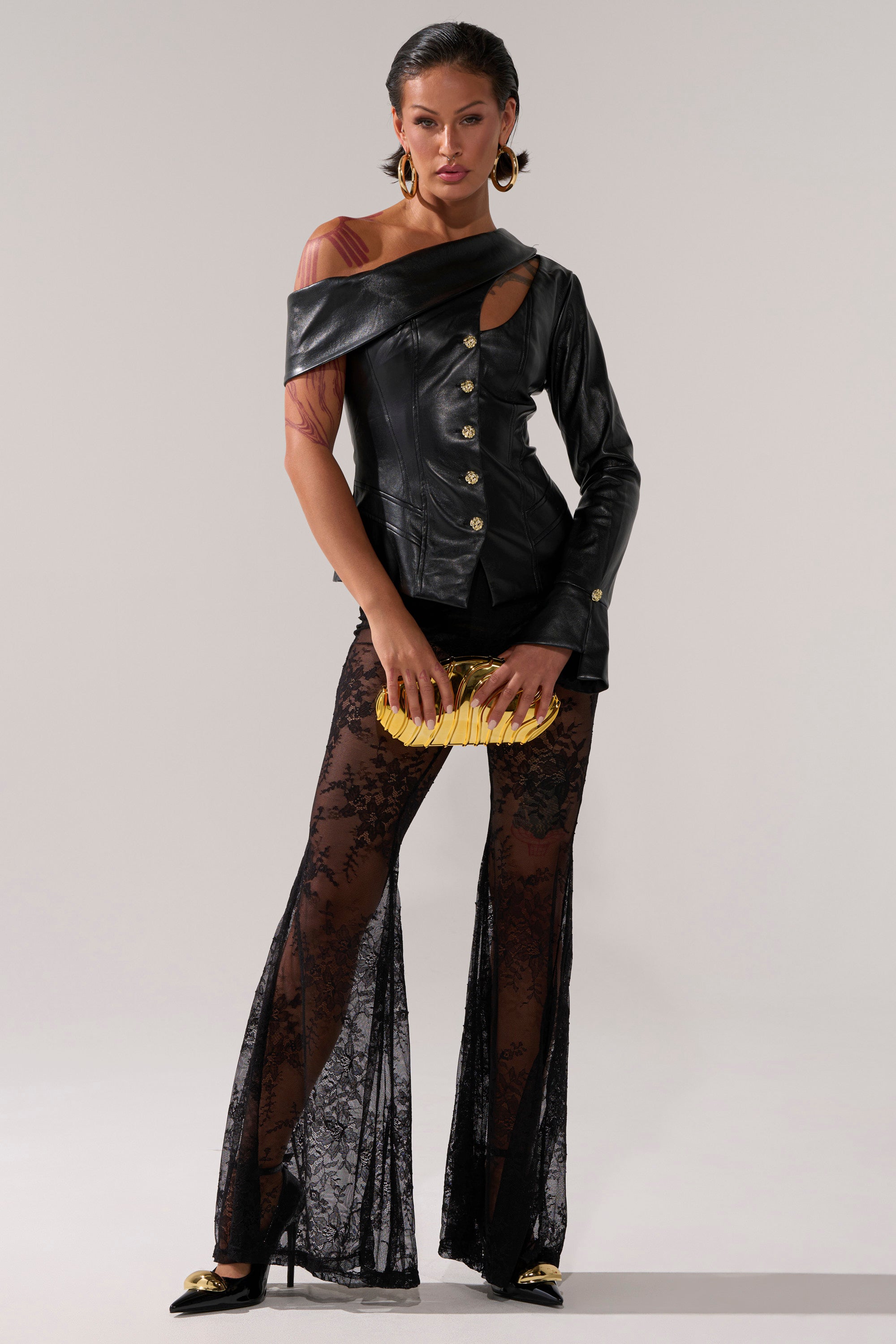 A woman poses in a chic look, wearing the YOU GOT ME FEELING FAUX LEATHER BUTTON DOWN with an asymmetrical neckline, sheer black lace pants, gold-tipped black heels, and a gold clutch against a plain light background.