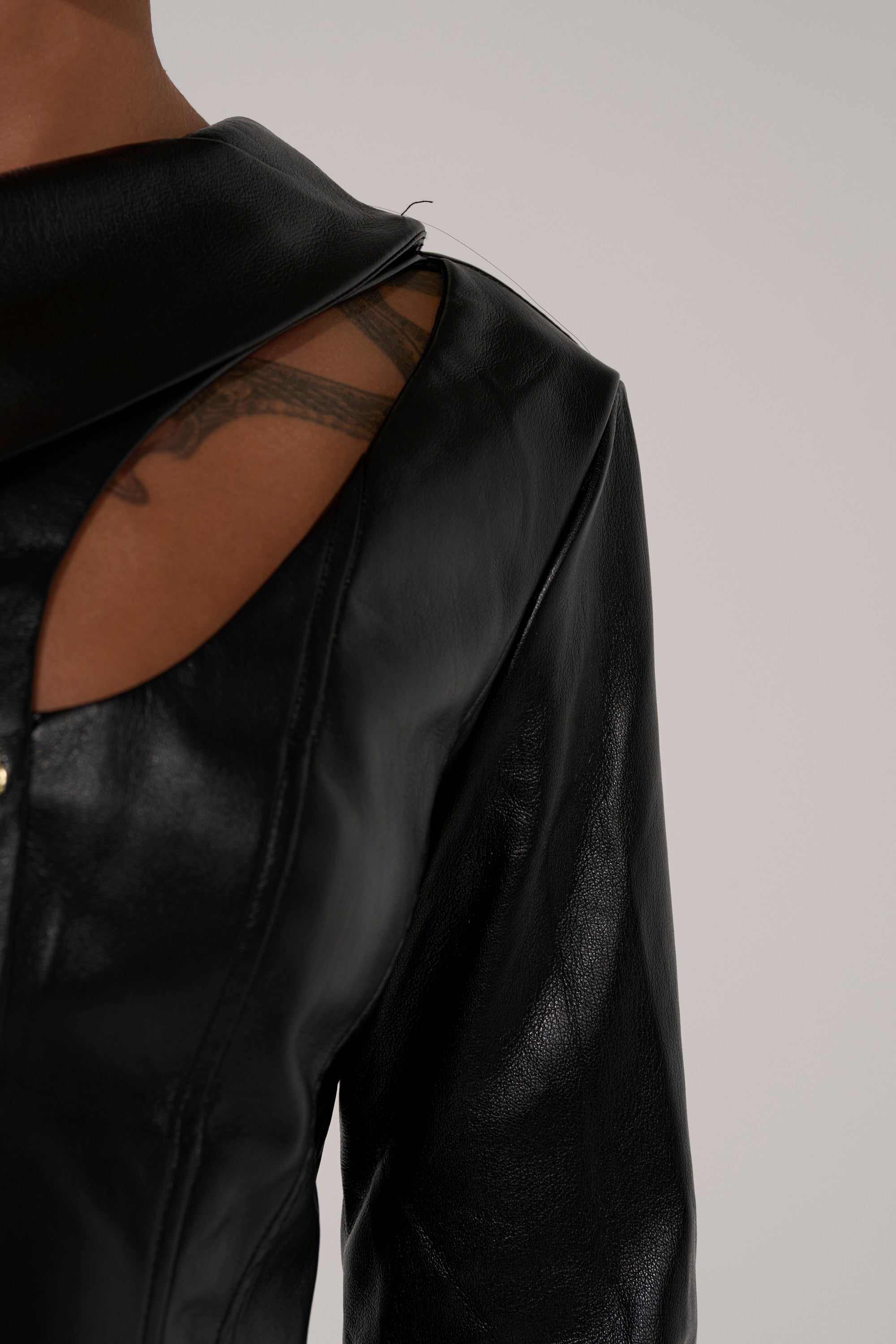 A close-up of a person wearing the YOU GOT ME FEELING FAUX LEATHER BUTTON DOWN, showcasing the shiny black material and an asymmetrical neckline that reveals part of a shoulder and a patterned tattoo against a plain light background.