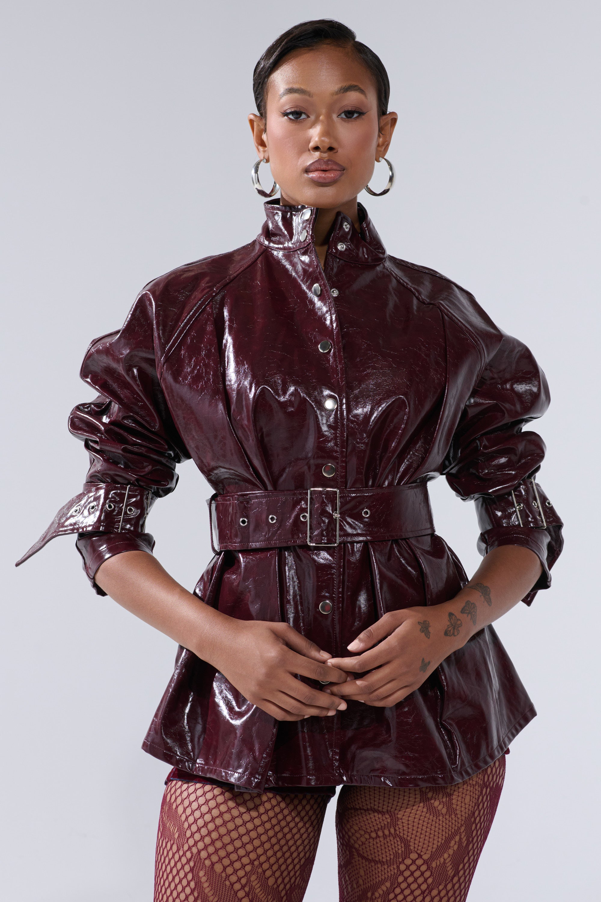 A woman poses confidently in the ON FIRE FAUX LEATHER BUTTON DOWN—a shiny burgundy patent jacket with an adjustable waist belt and puffed sleeves—paired with fishnet stockings, hoop earrings, and a slicked-back hairstyle against a plain light background.