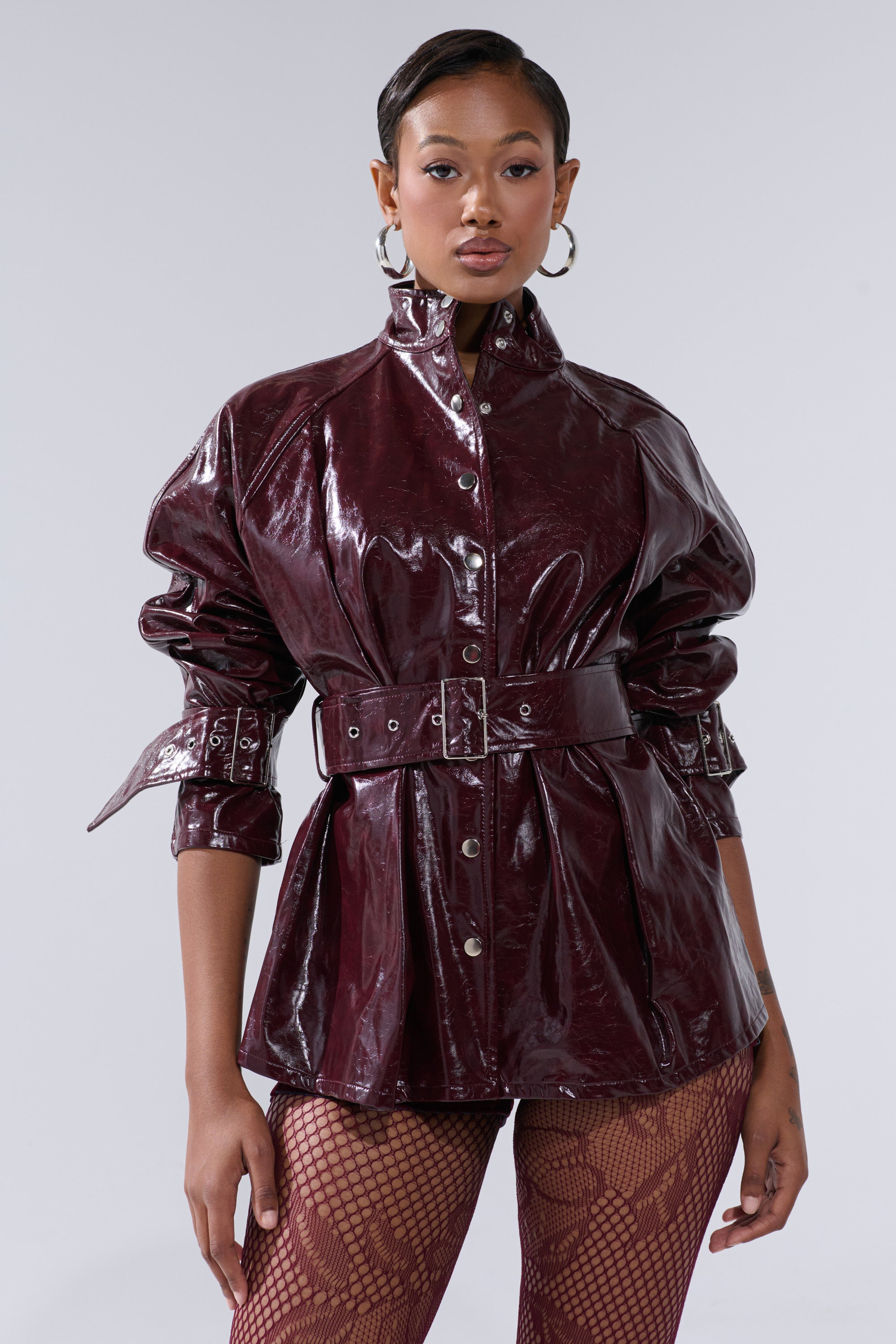 A person stands confidently in the ON FIRE FAUX LEATHER BUTTON DOWN, a shiny dark burgundy belted jacket with silver buttons. Slicked-back hair, hoop earrings, and fishnet tights complete the look against a plain light gray background.