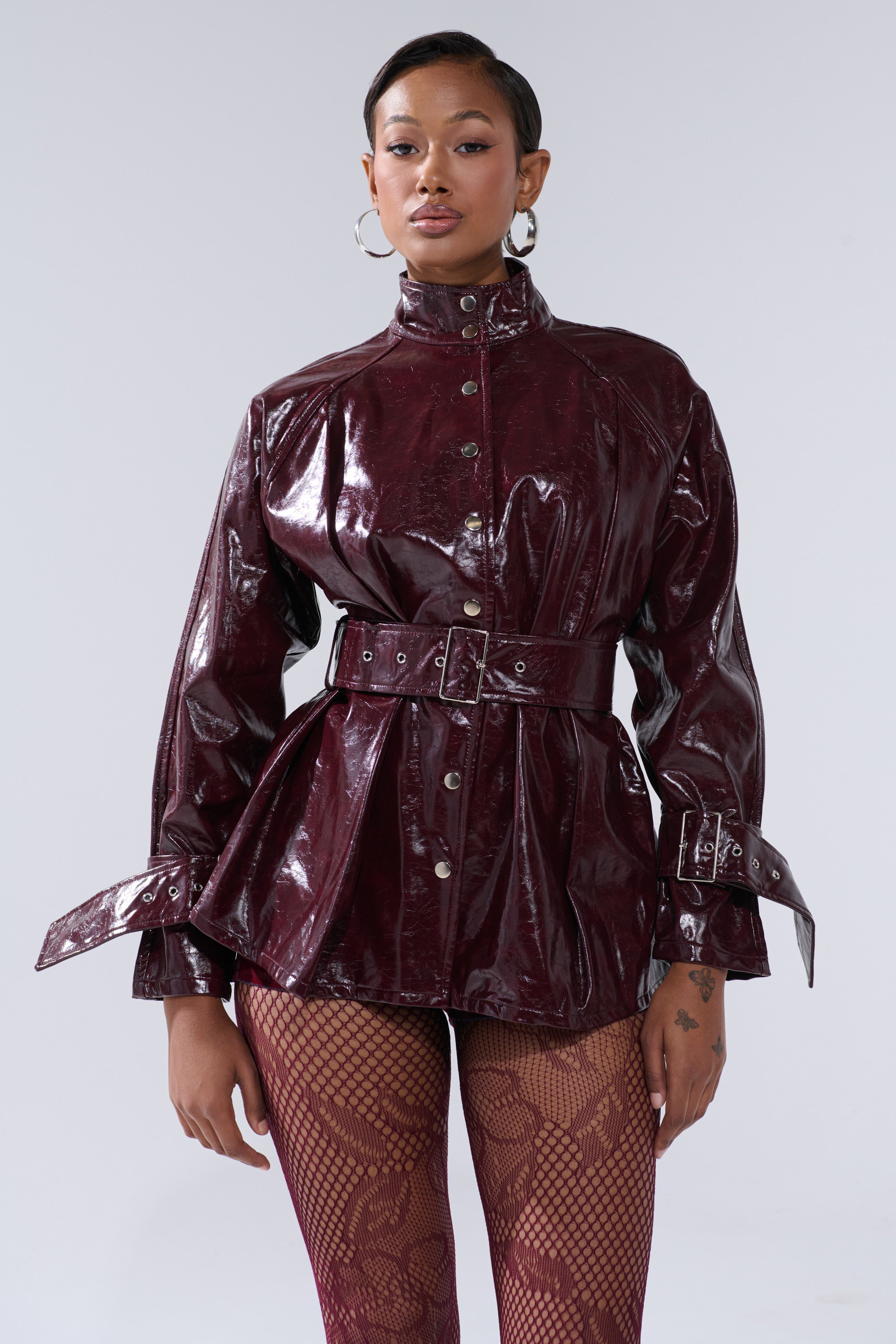 A woman wears the ON FIRE FAUX LEATHER BUTTON DOWN jacket with button details and maroon patterned tights, standing against a plain light background.