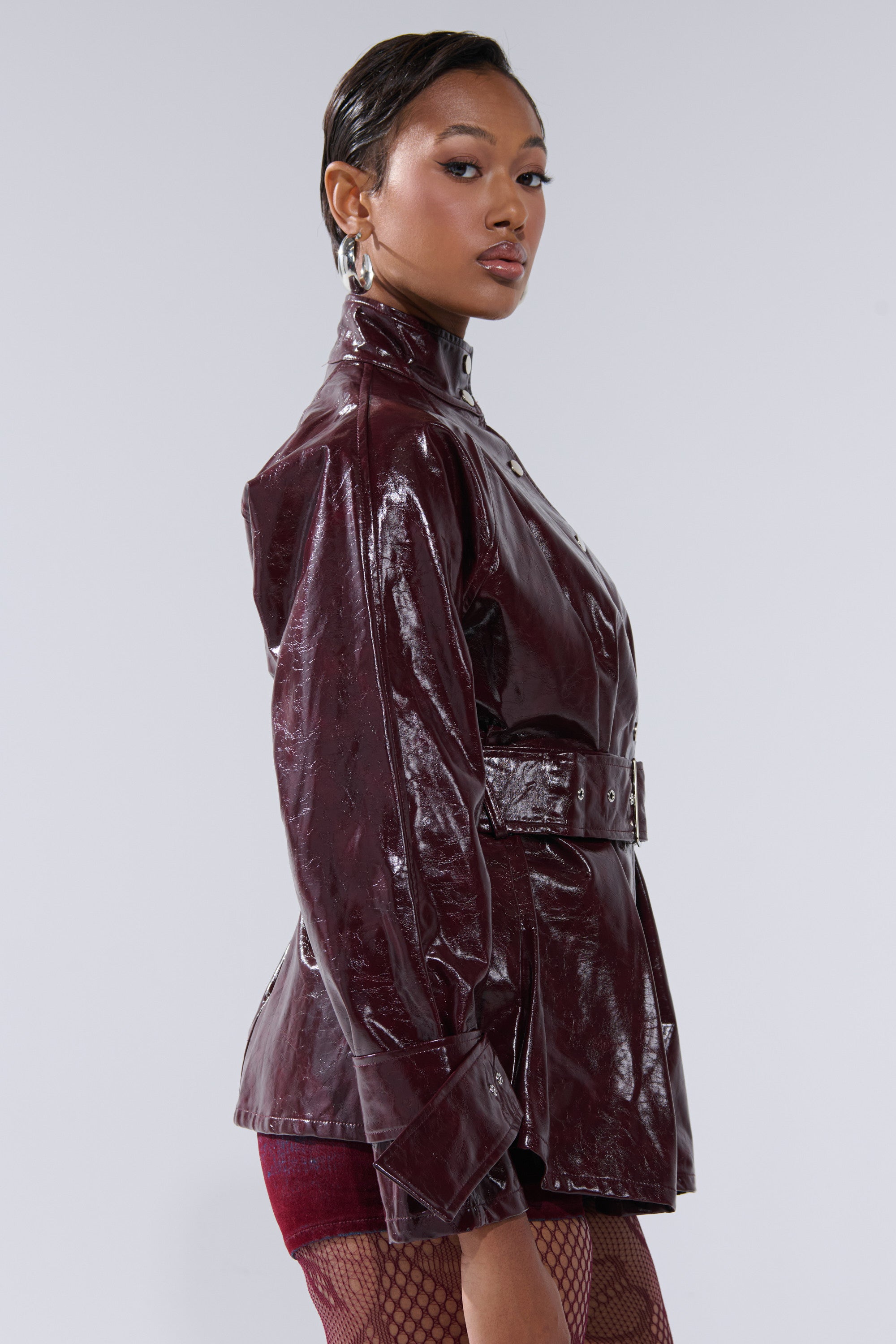 A woman in profile wears the ON FIRE FAUX LEATHER BUTTON DOWN, a shiny maroon belted jacket, paired with hoop earrings and fishnet stockings. Her hair is slicked back against a neutral background.