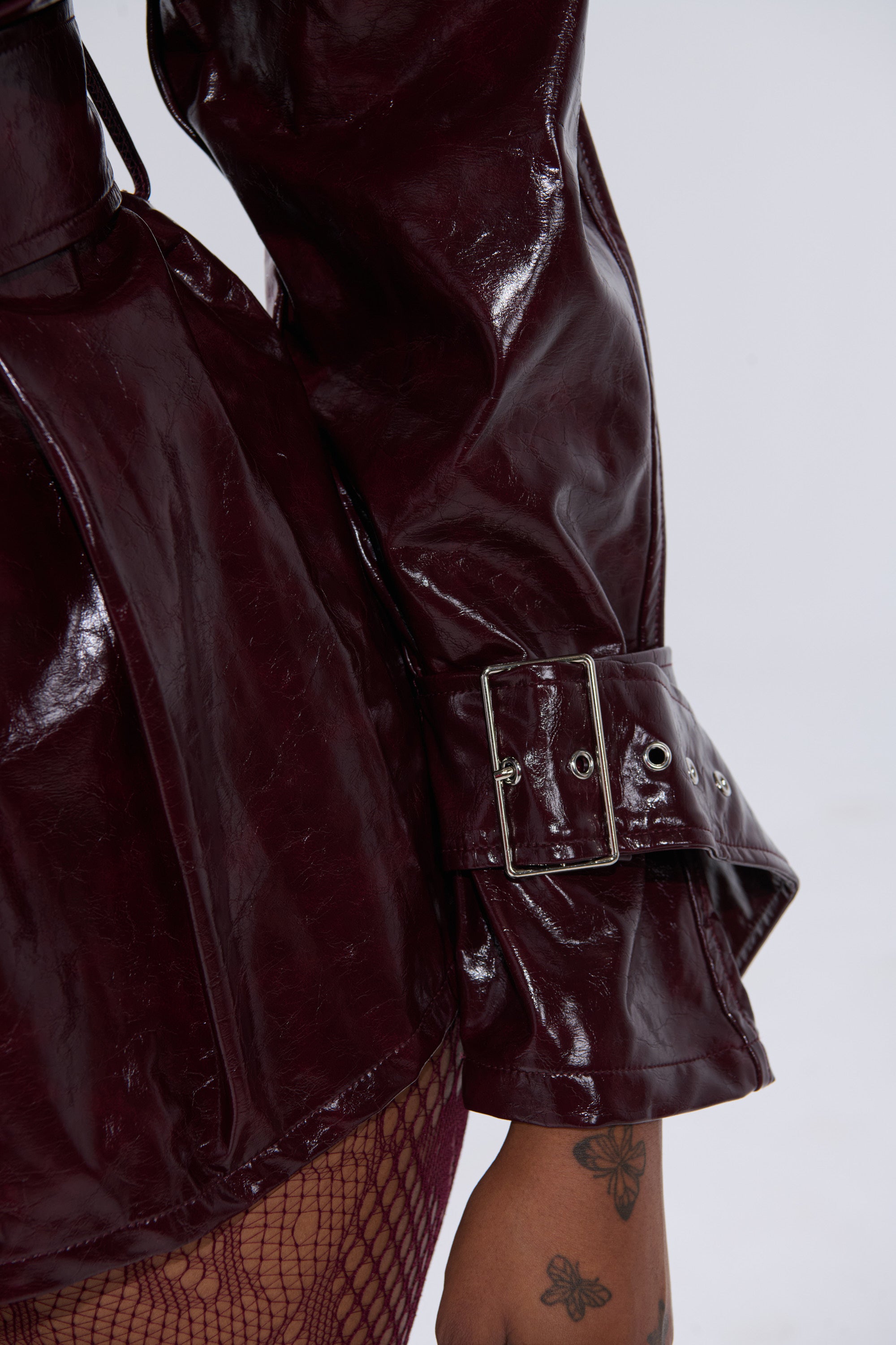 A close-up of a person wearing the ON FIRE FAUX LEATHER BUTTON DOWN in shiny burgundy patent faux leather, featuring an adjustable waist belt and belted sleeves. Their tattooed hand rests at their side, with fishnet stockings partially visible.