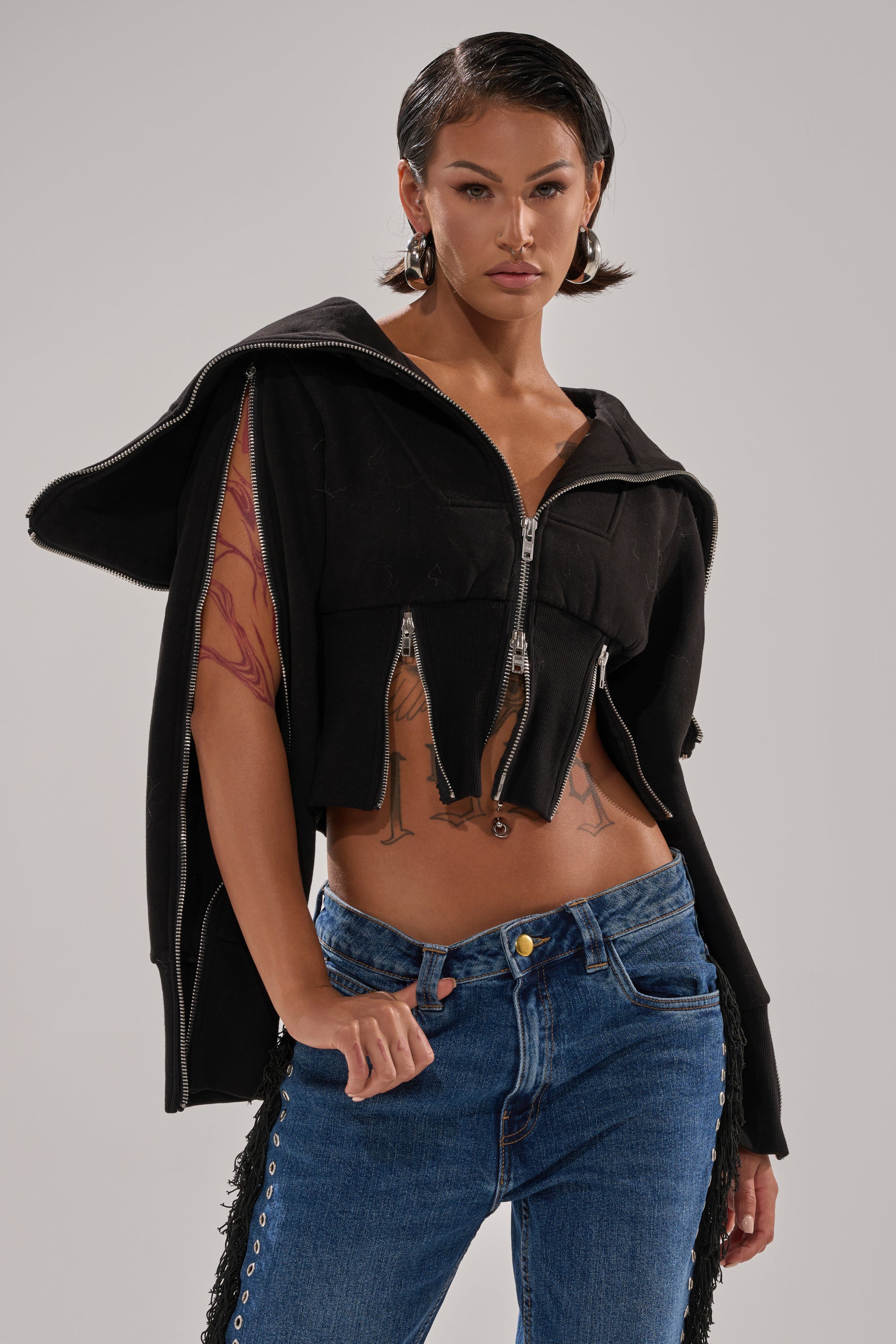 A person with short, styled hair confidently models the MULTI ZIP CORSETED HOODIE—a black cropped jacket with zip accents—paired with blue jeans. Tattooed arms are partially visible against a plain, light background. 0