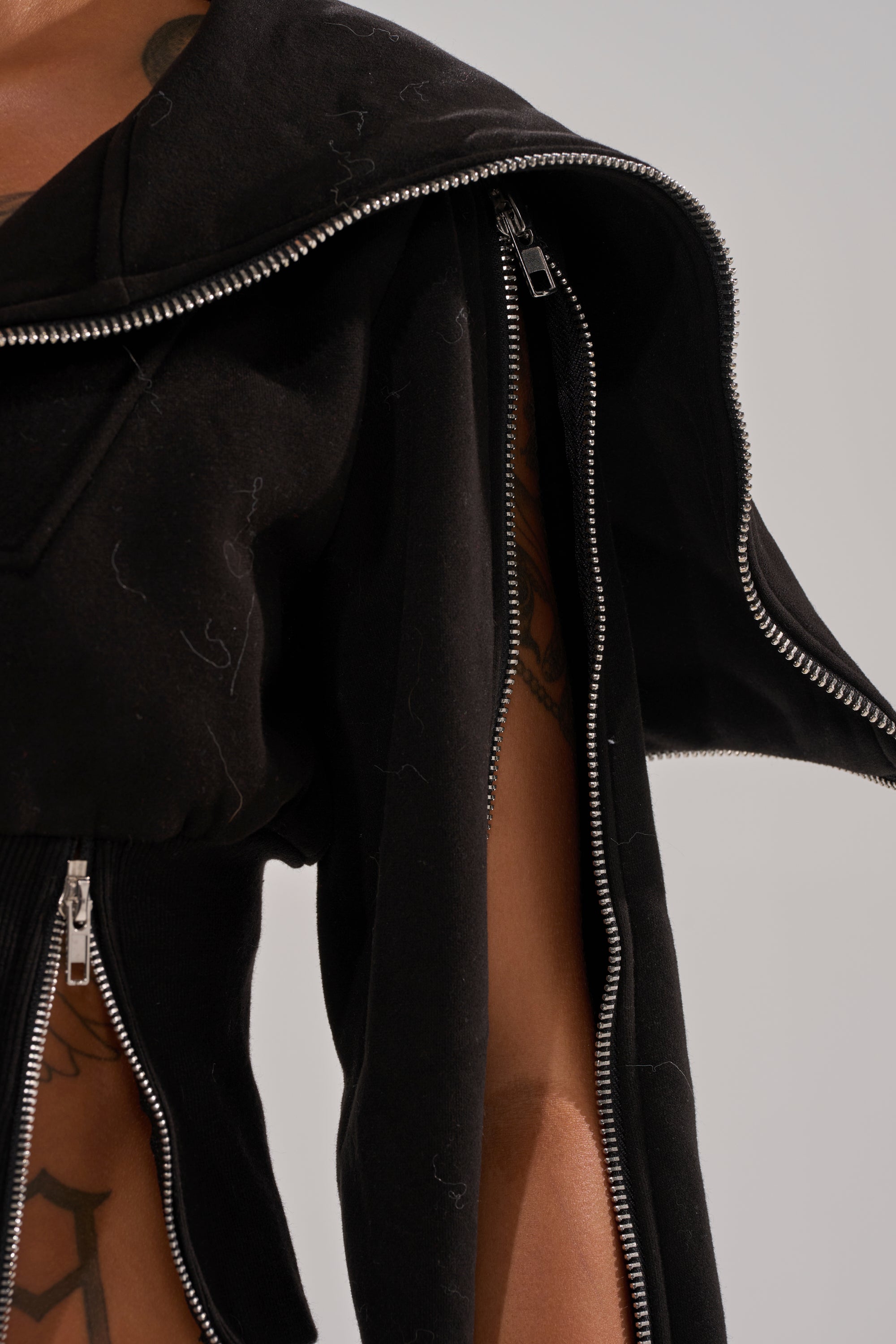 A close-up of a person wearing the MULTI ZIP CORSETED HOODIE, a black cropped sweatshirt with bold silver zippers. The partially open multi-zip design reveals skin and tattoos, creating an edgy and unconventional look. 9