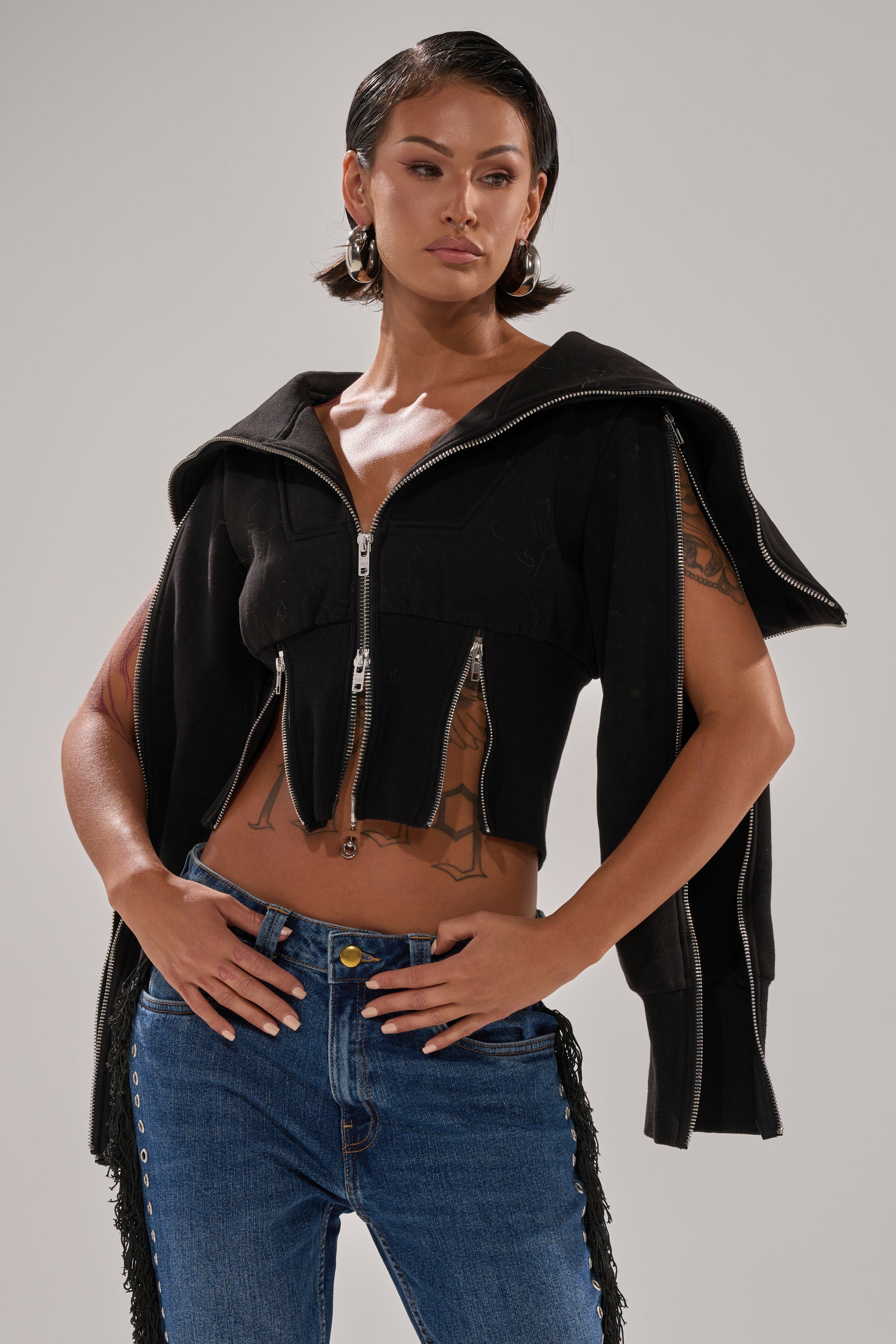 A woman with short dark hair and hoop earrings poses confidently in the MULTI ZIP CORSETED HOODIE, featuring zip details and cut-off sleeves that reveal a tattooed arm, styled with blue jeans against a plain background. 2