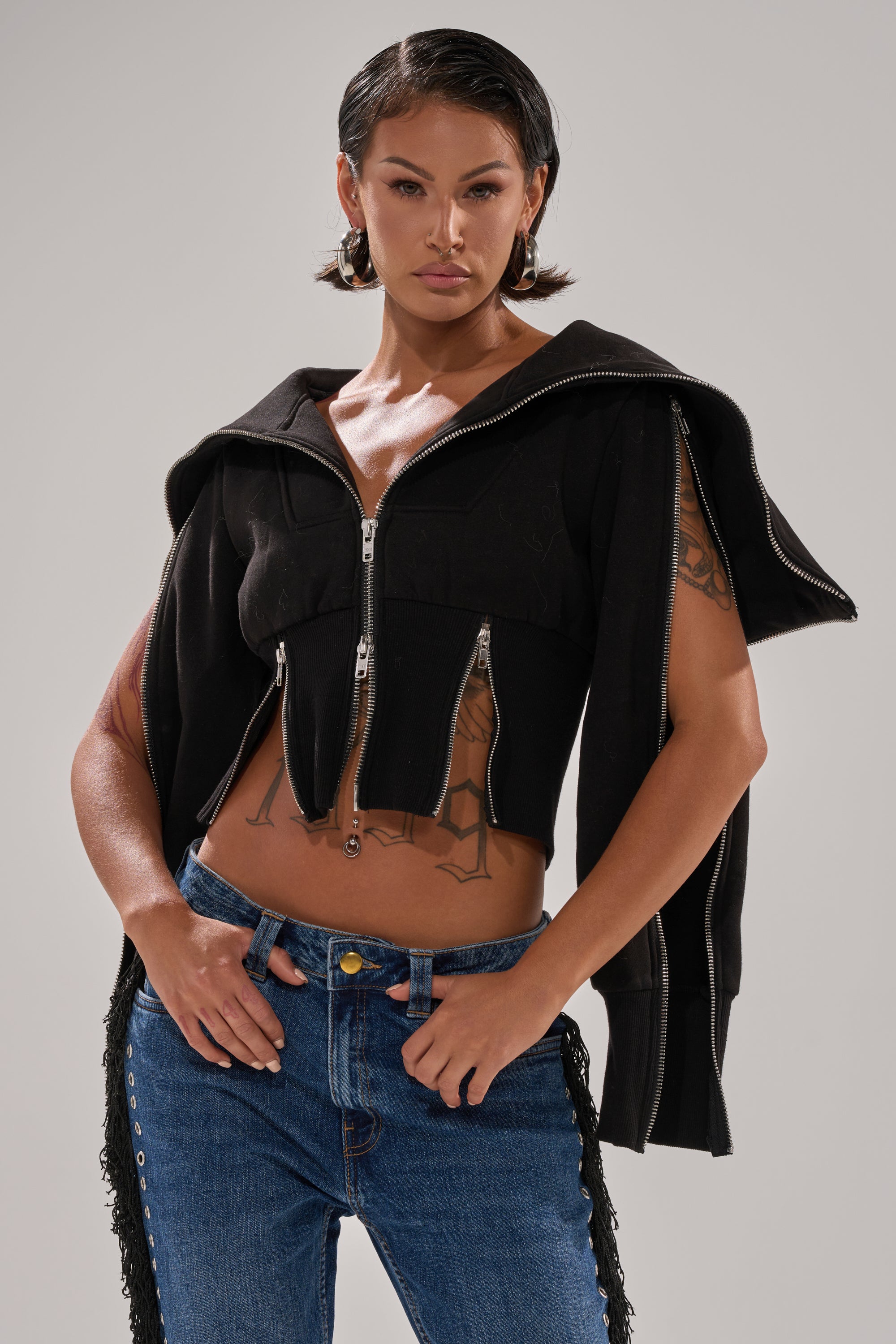A woman with short dark hair poses confidently in the MULTI ZIP CORSETED HOODIE draped over her shoulders, showcasing her midriff and tattoos. She pairs it with blue jeans and hoop earrings against a plain background. 3