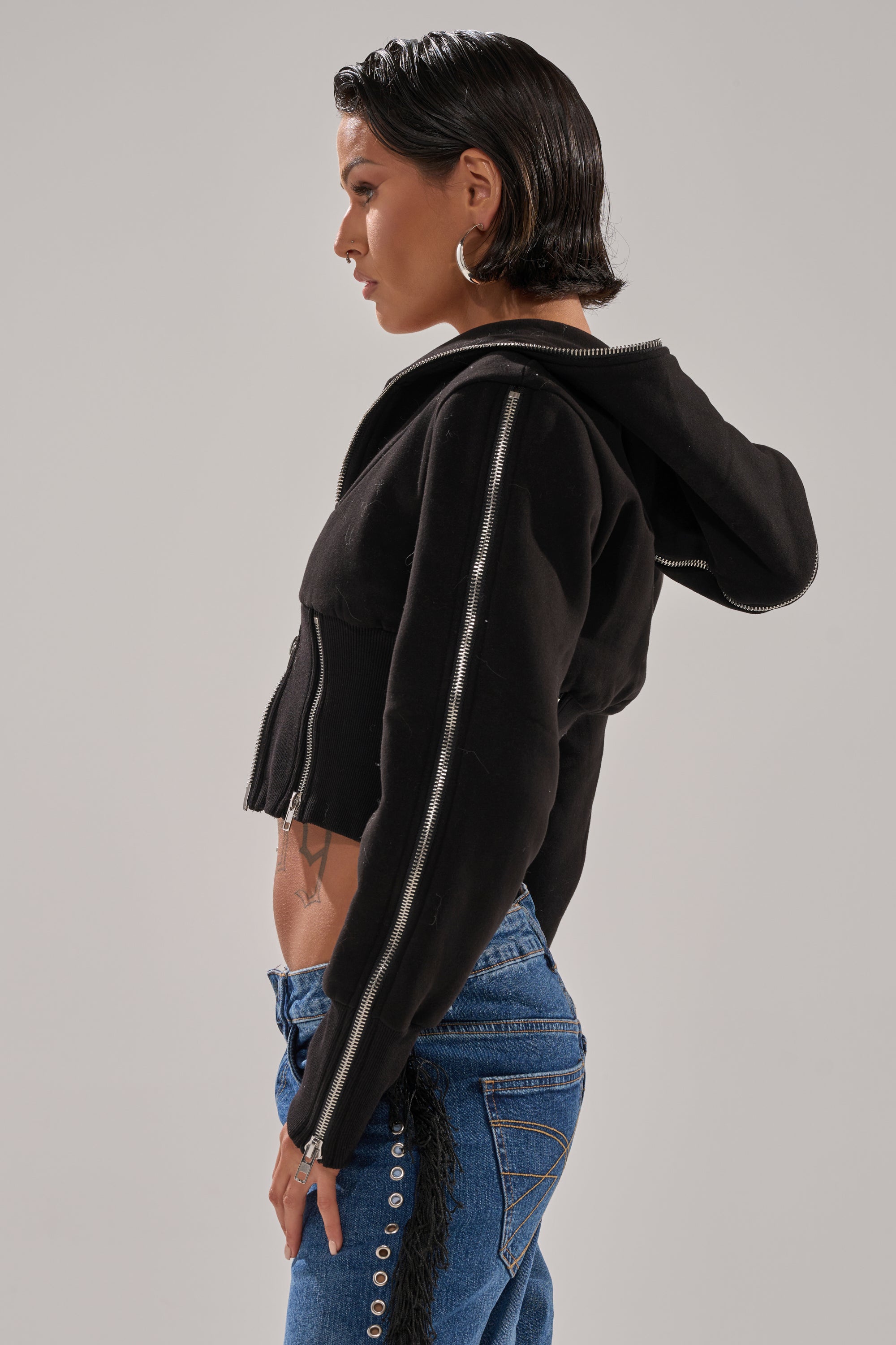 A person with short dark hair and hoop earrings poses in profile wearing the MULTI ZIP CORSETED HOODIE, a black cropped sweatshirt, paired with blue jeans featuring decorative buttons and pocket details against a plain background. 4