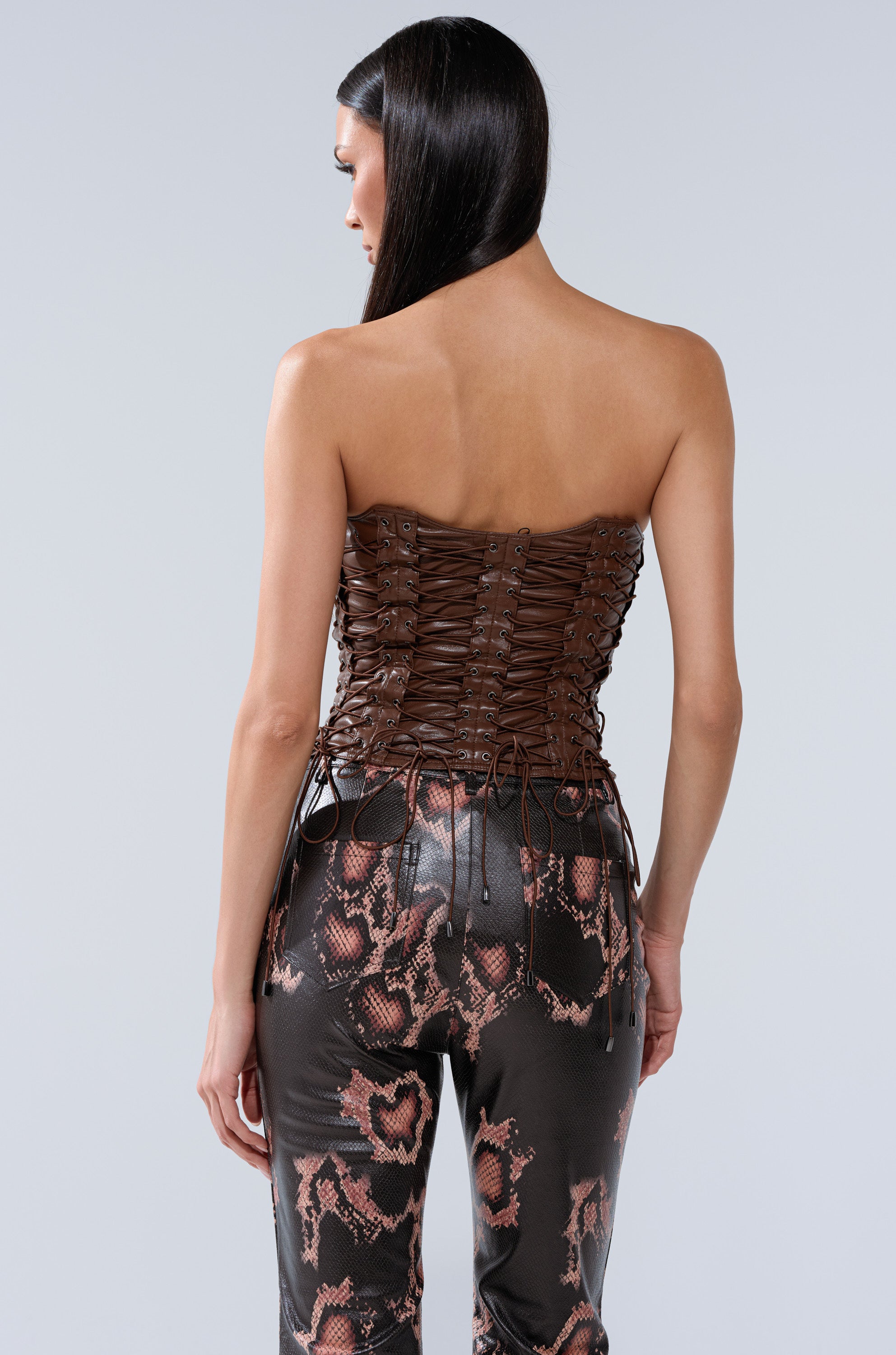 A woman with straight dark hair faces away, wearing the KNOTTED UP FAUX LEATHER CORSET in brown and high-waisted black pants with a pink snake print. The setting is against a light gray background.