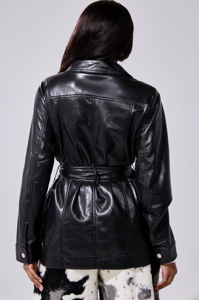 A person with long dark hair is seen from the back, wearing the TALK TO ME BABY FAUX LEATHER BUTTON DOWN—an oversized black jacket with an adjustable waist belt—and black-and-white patterned pants, against a plain light background.