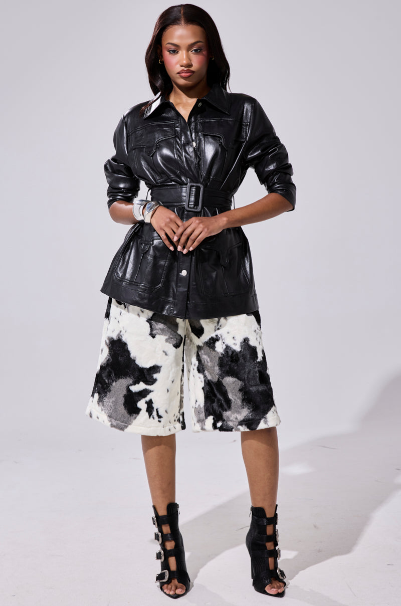 A woman stands confidently in the TALK TO ME BABY FAUX LEATHER BUTTON DOWN, paired with wide black-and-white cow-print shorts and black cut-out high-heeled sandals, set against a plain light background.