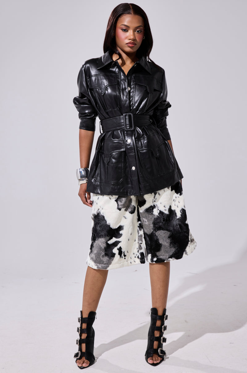 A woman stands confidently in the TALK TO ME BABY FAUX LEATHER BUTTON DOWN, paired with a black and white cow-print knee-length skirt, black high-heeled buckled sandals, and a large silver cuff bracelet.