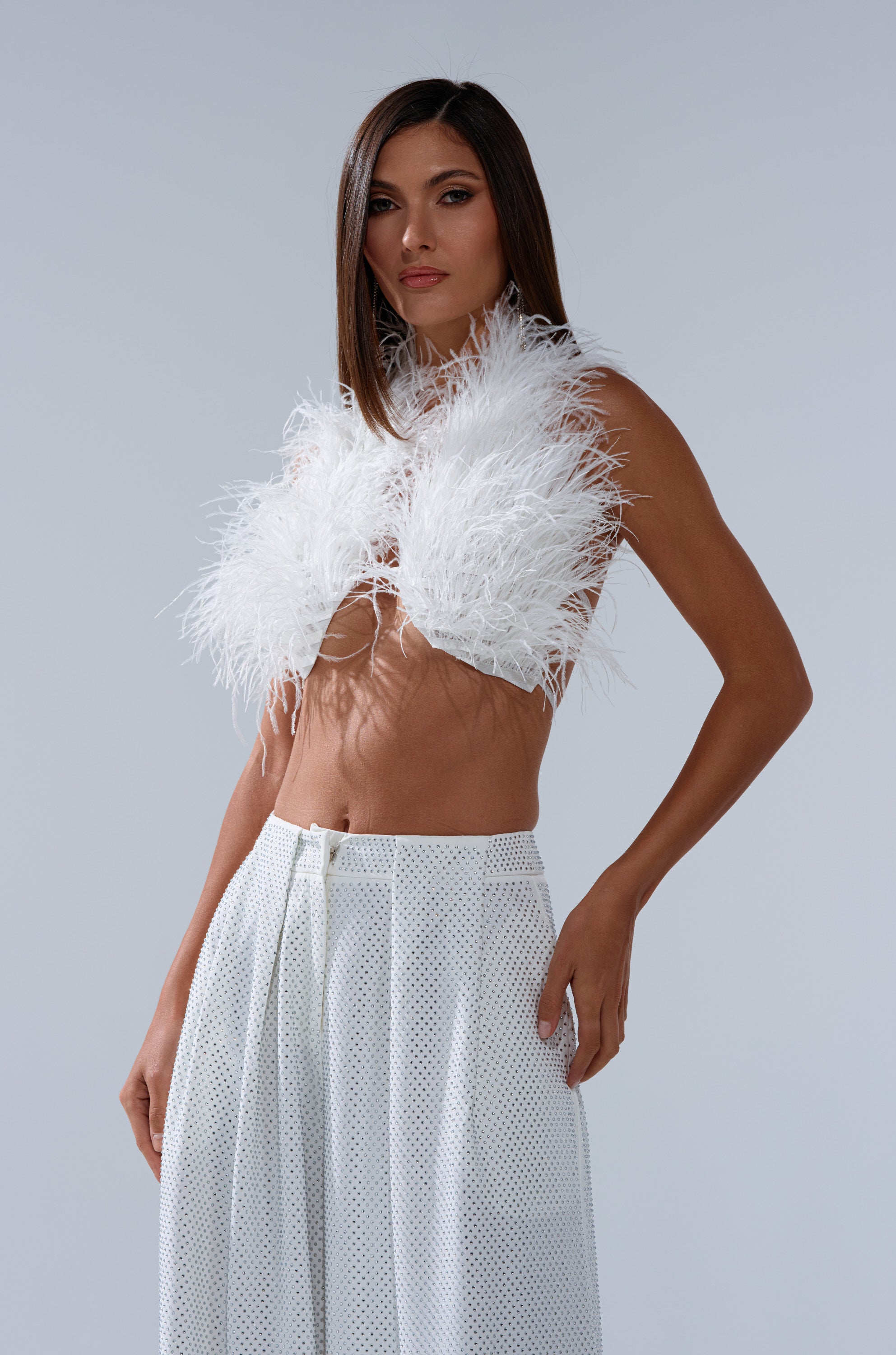 A woman poses confidently against a plain background in the SHAKE IT FOR ME GIRL FEATHER BRALETTE and high-waisted white pants, her long straight hair down and one hand on her hip. 1