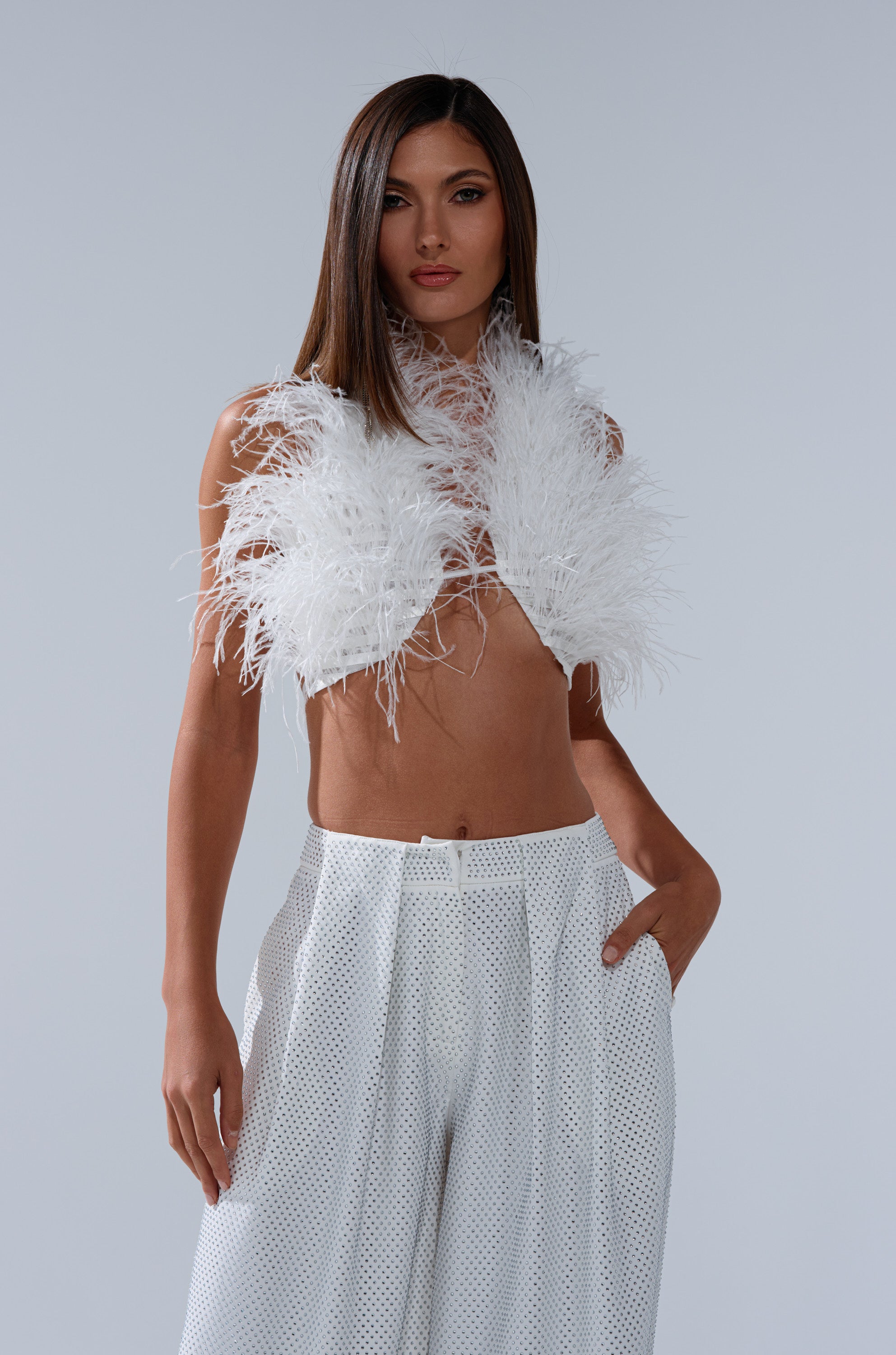A woman poses against a plain background wearing the SHAKE IT FOR ME GIRL FEATHER BRALETTE and high-waisted, wide-leg white pants. Her long, straight hair is worn down as she rests her hand in her pocket. 2