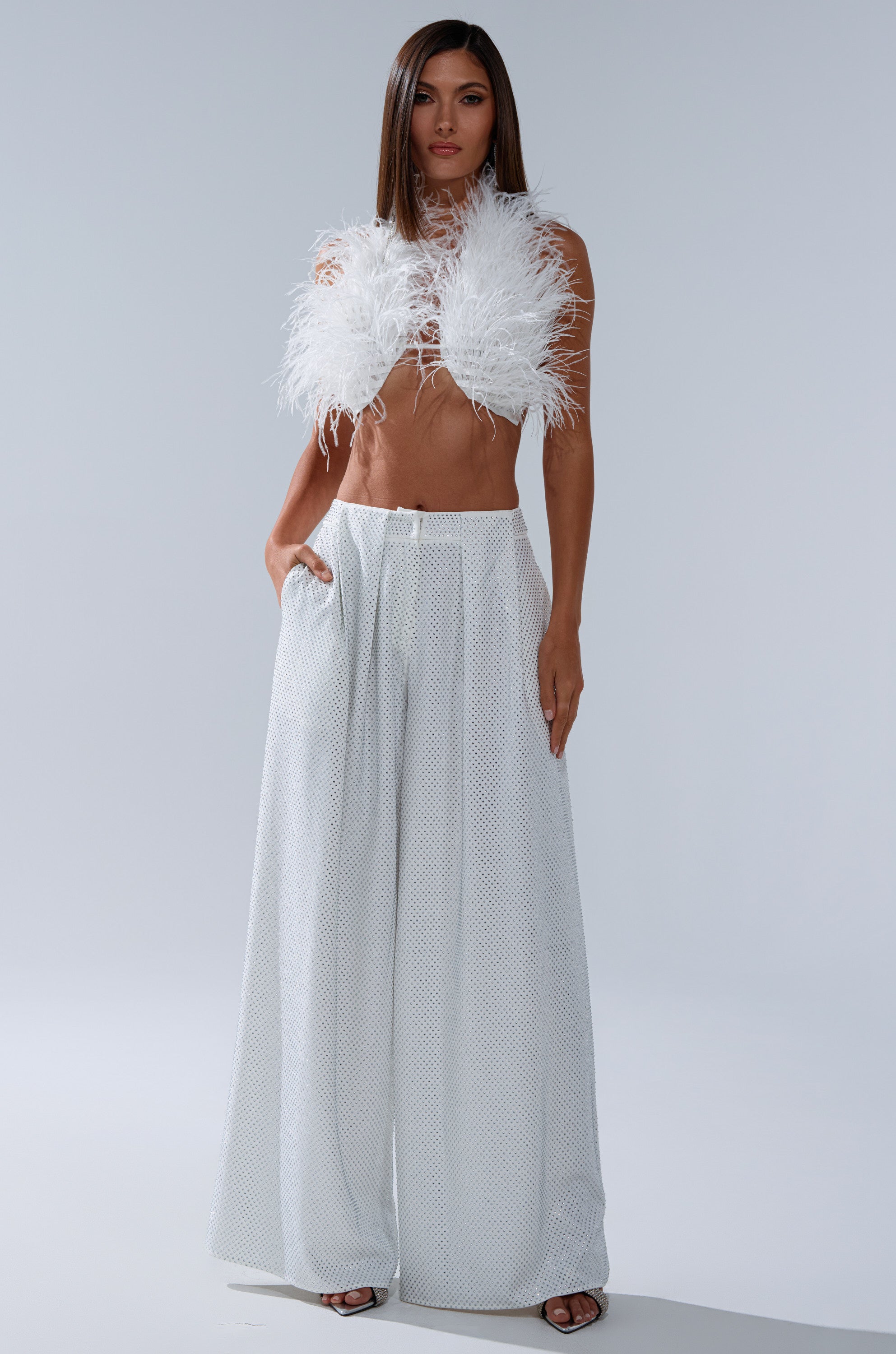A woman poses against a plain light background in high-waisted wide-leg white pants and the SHAKE IT FOR ME GIRL FEATHER BRALETTE, with her hands in her pockets. 6