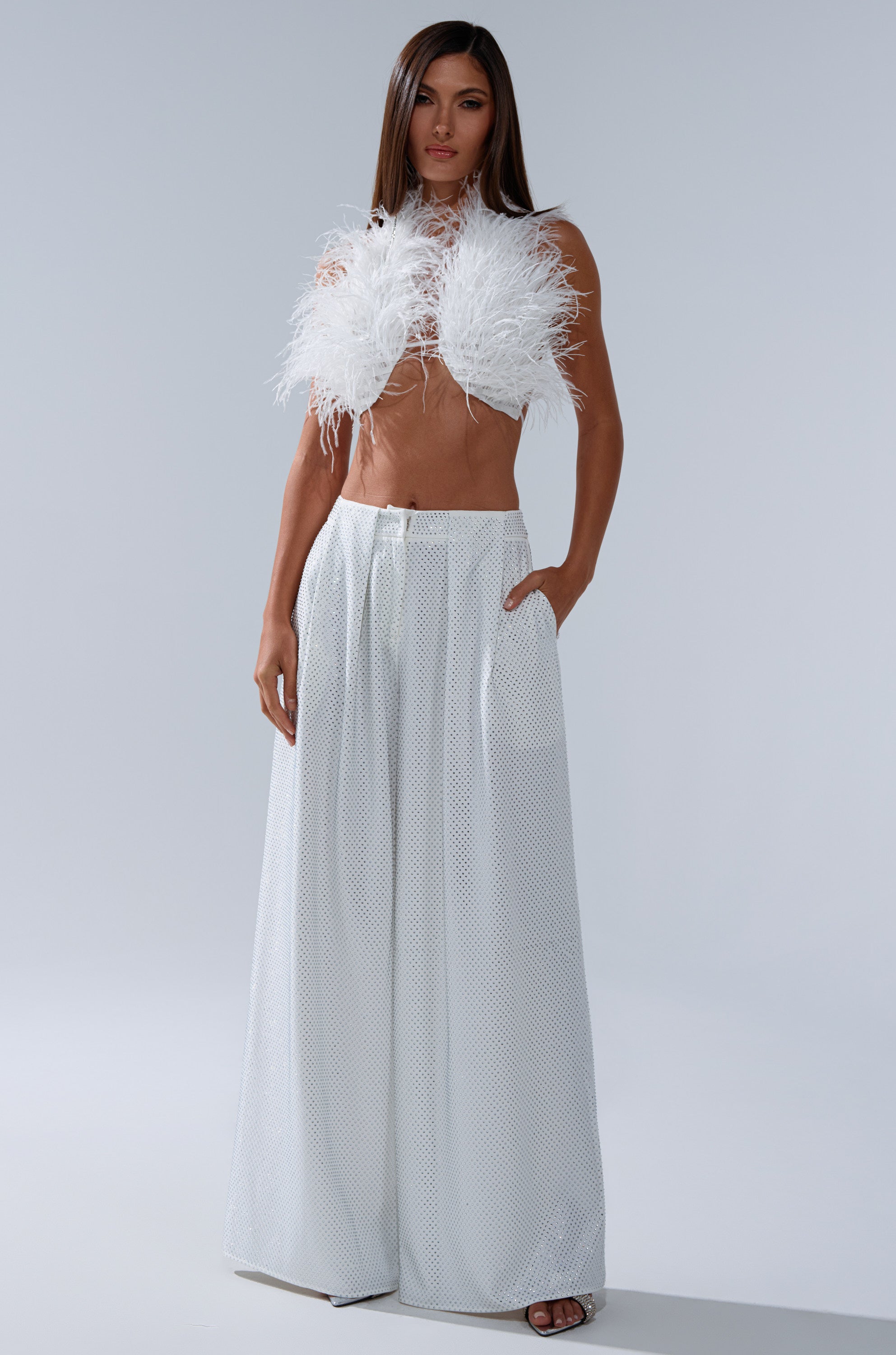 A woman stands against a light gray background, wearing the SHAKE IT FOR ME GIRL FEATHER BRALETTE in white and high-waisted, wide-leg white pants, with her hands in her pockets. 7