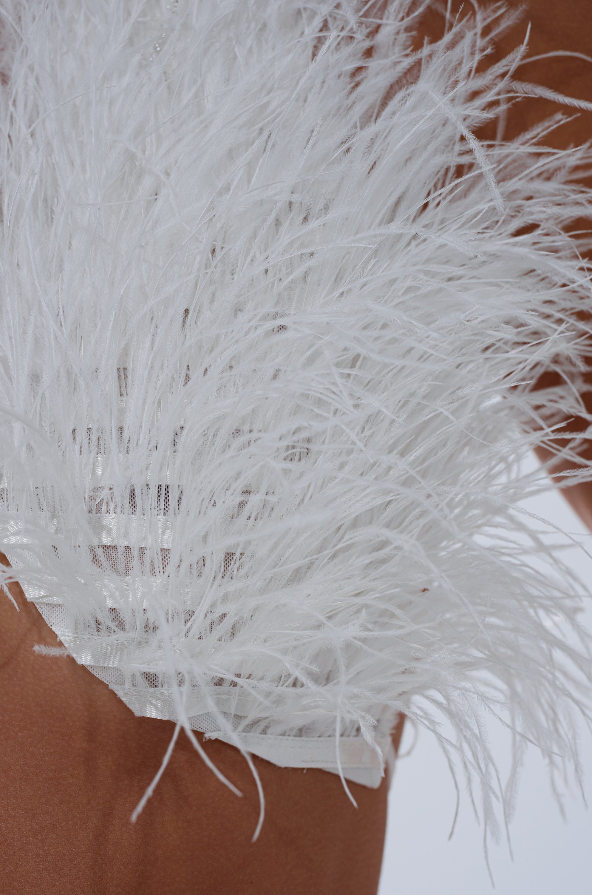 Close-up of the SHAKE IT FOR ME GIRL FEATHER BRALETTE, featuring white feathery trim fanning out along sheer mesh, creating striking contrast against the smooth, brown fabric background. 8