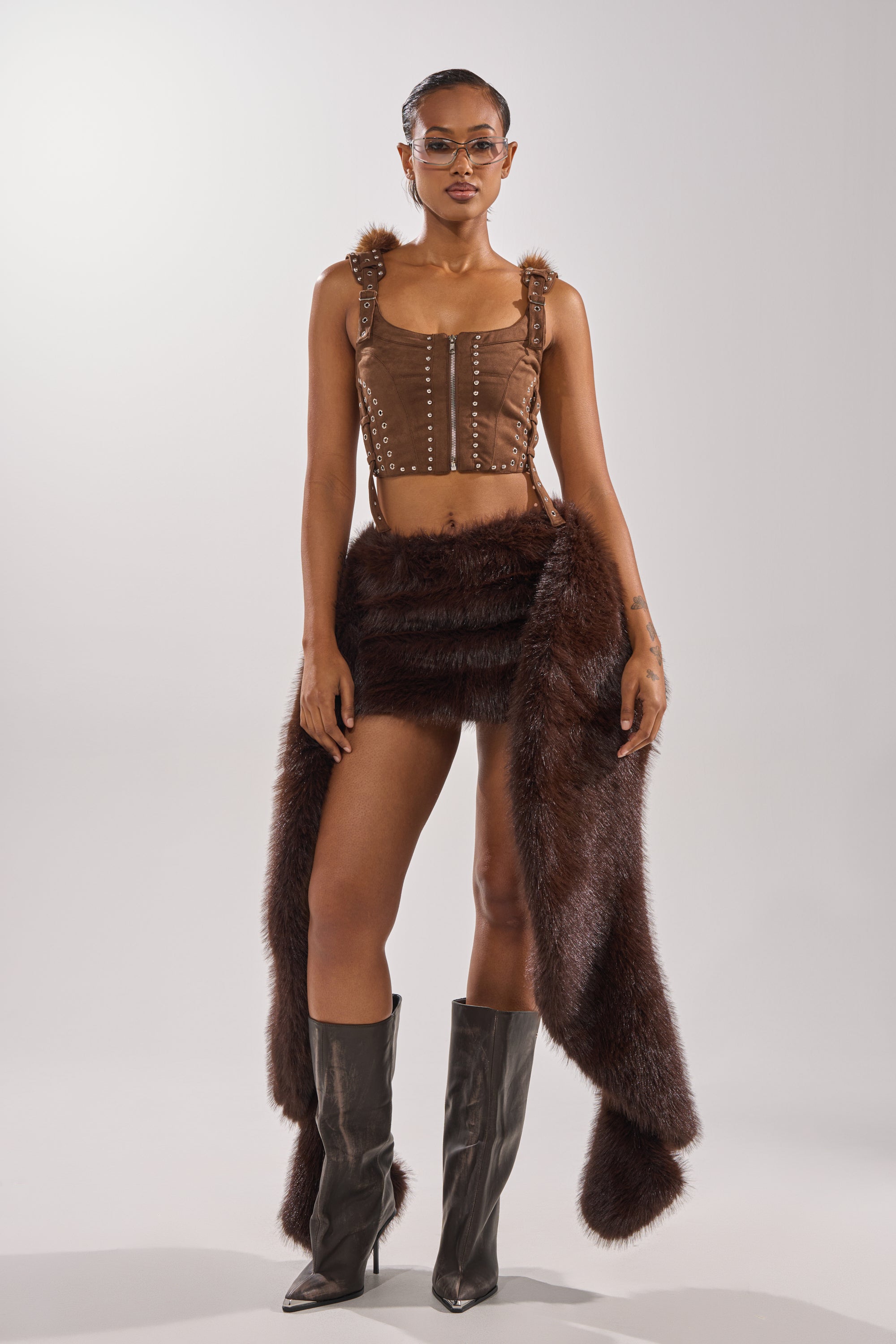 A woman stands confidently in the TAKE ME TO THE RODEO HOODED FUR TOP, paired with a faux fur mini skirt, sash, knee-high heeled boots, and clear glasses against a plain light background. 7
