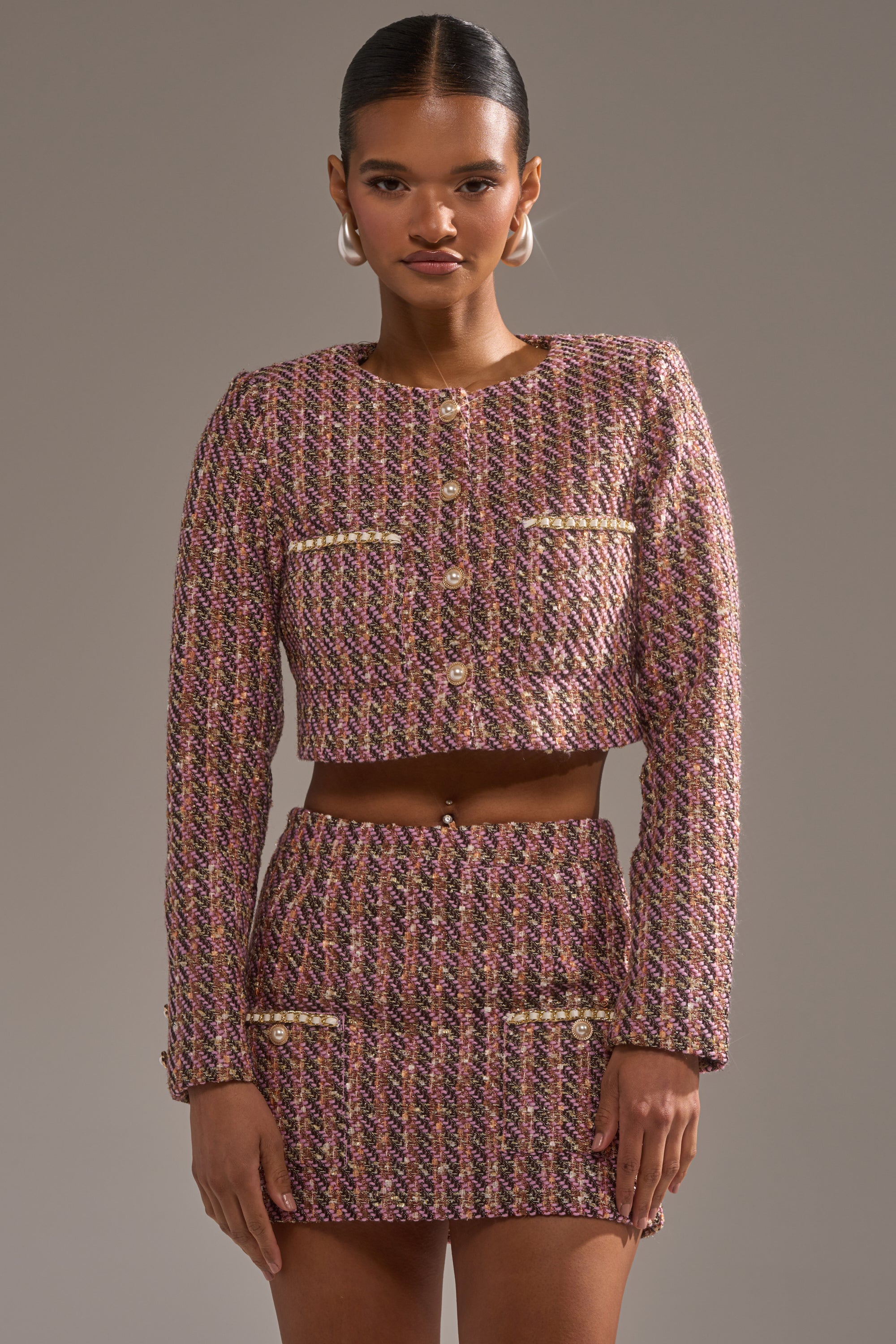 A woman wears the TRUTH OR DARE TWEED BLAZER—pink, gold, and brown with a cropped fit and gold buttons—paired with a matching high-waisted mini skirt with metallic pocket details, standing against a plain gray background. 0