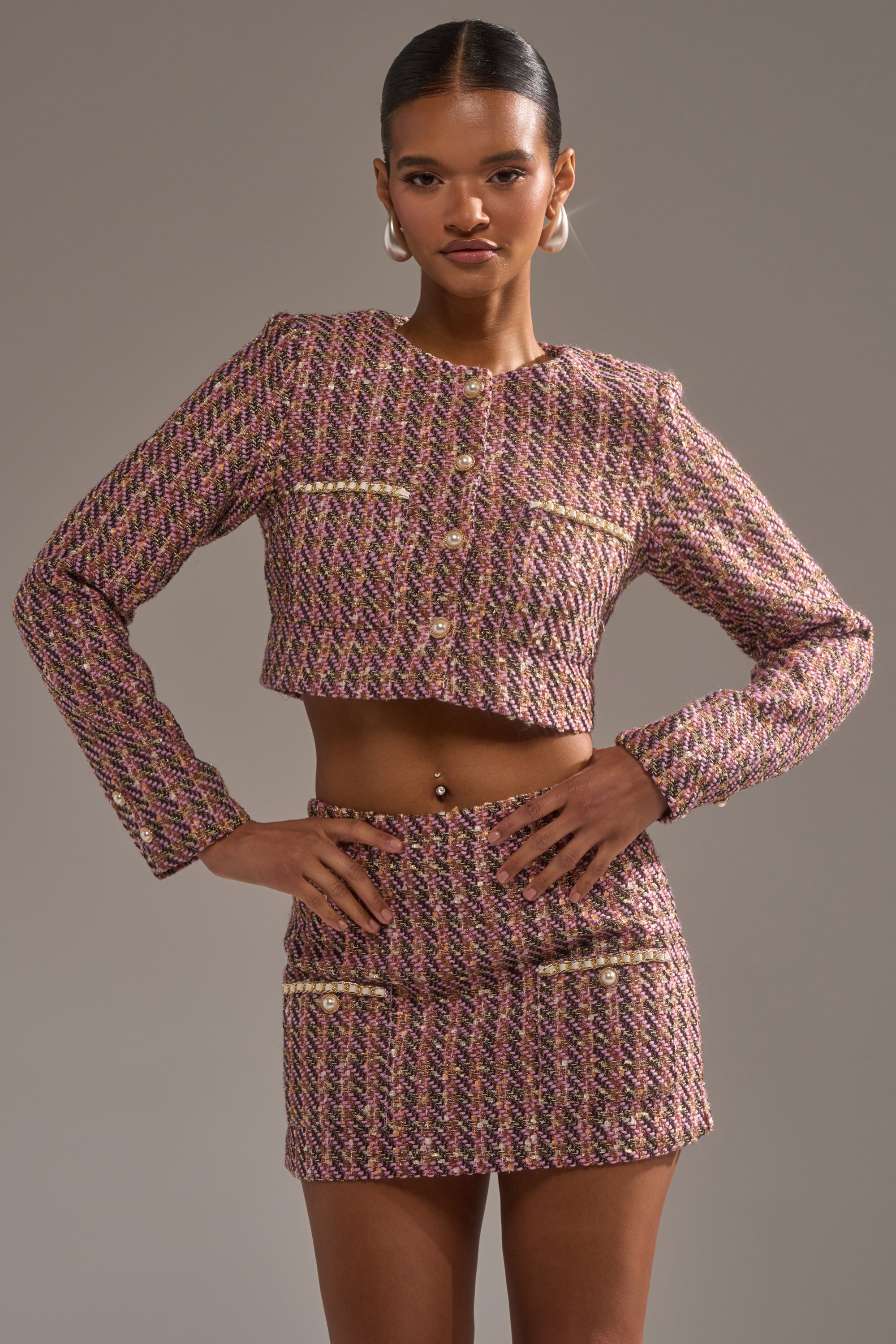 A woman poses confidently in the TRUTH OR DARE TWEED BLAZER, a textured, multicolored blazer with gold buttons, paired with a matching high-waisted mini skirt. She accessorizes with hoop earrings and places her hands on her hips. 1