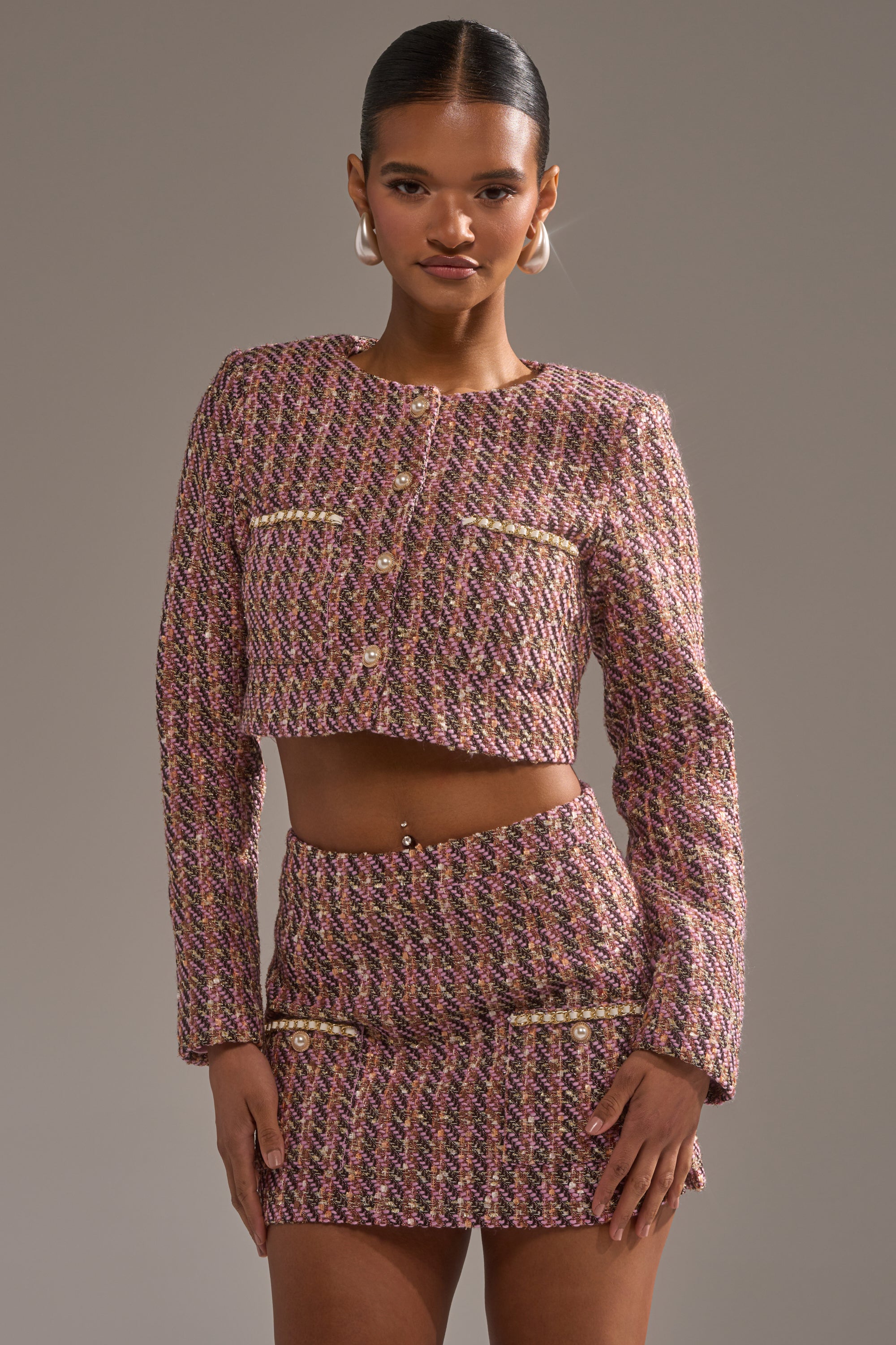 A woman poses confidently in the TRUTH OR DARE TWEED BLAZER with a cropped fit and matching high-waisted mini skirt with gold buttons, standing against a plain gray background. 2