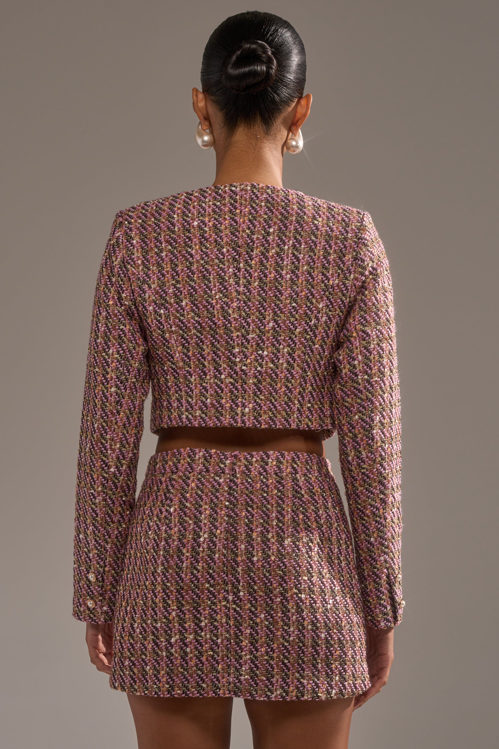 A woman with a sleek bun and large pearl earrings wears the TRUTH OR DARE TWEED BLAZER in a multicolored cropped fit, paired with a matching high-waisted mini skirt, standing against a plain grey background. 4