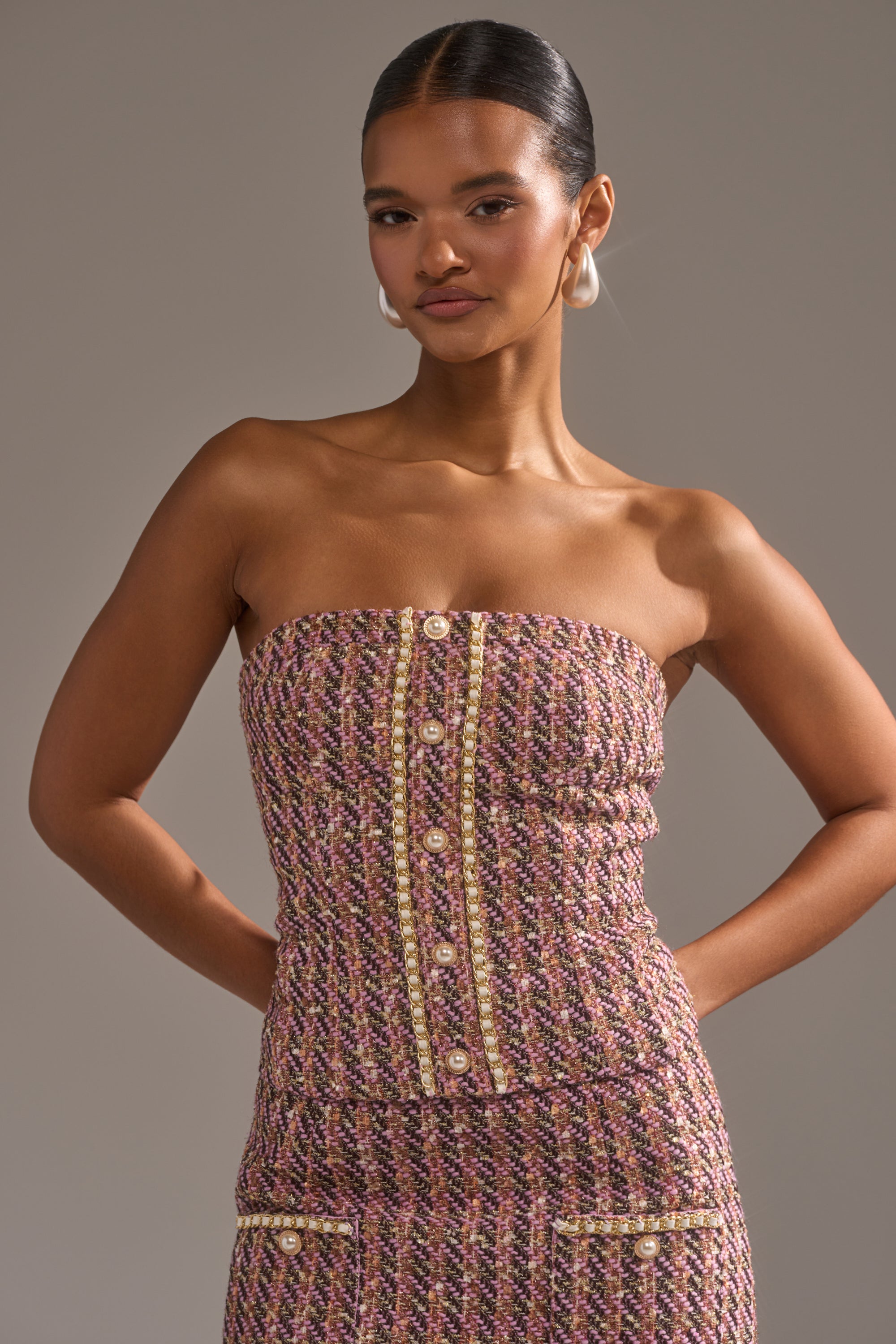 A woman with slicked-back hair poses with her hands behind her back, wearing the TRUTH OR DARE TWEED CORSET in textured pink and gold, paired with pearl hoop earrings, against a plain taupe background. 0