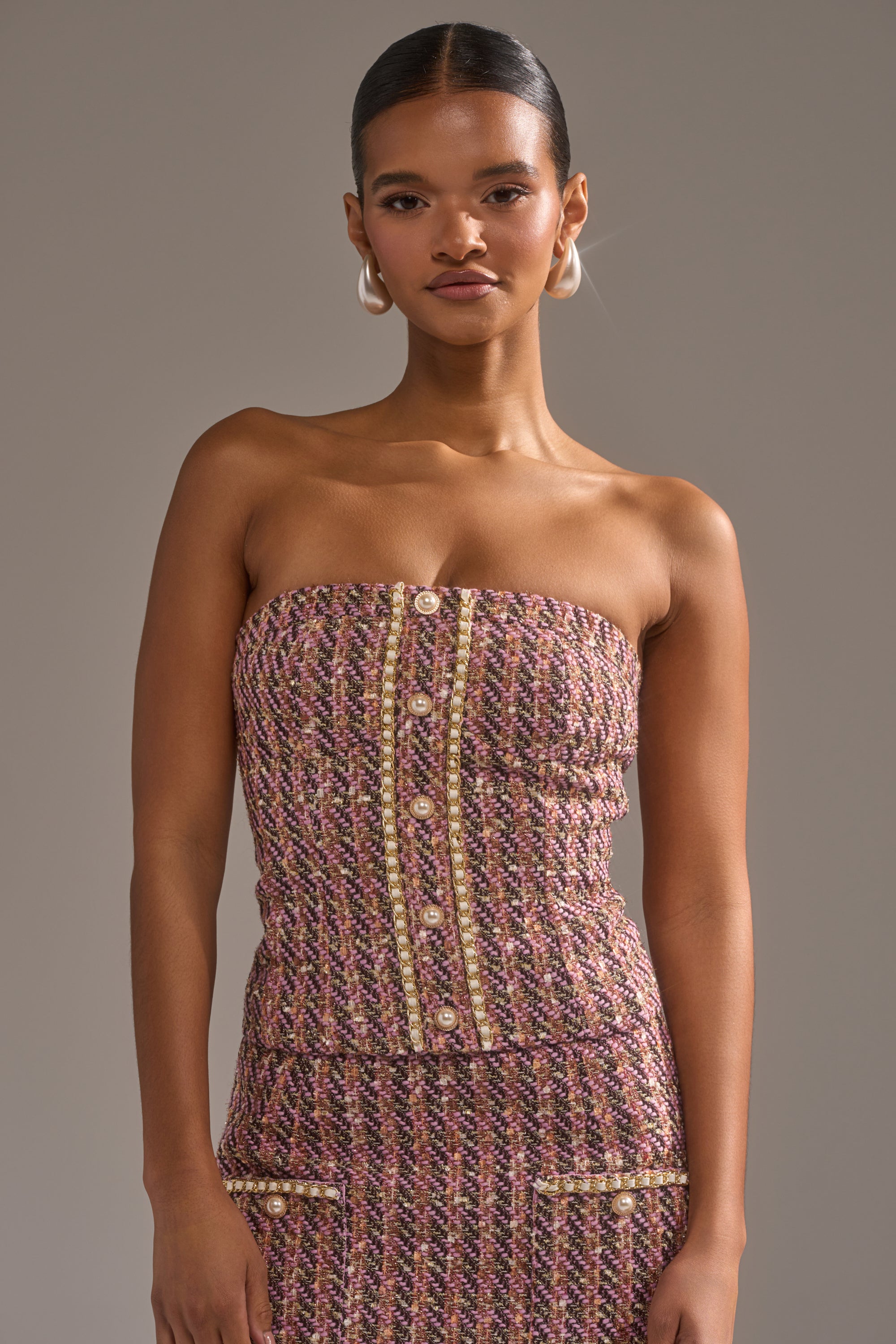 A woman with slicked-back hair wears the TRUTH OR DARE TWEED CORSET, a strapless mauve and gold button-up top paired with a matching skirt, standing confidently against a plain gray background. 1