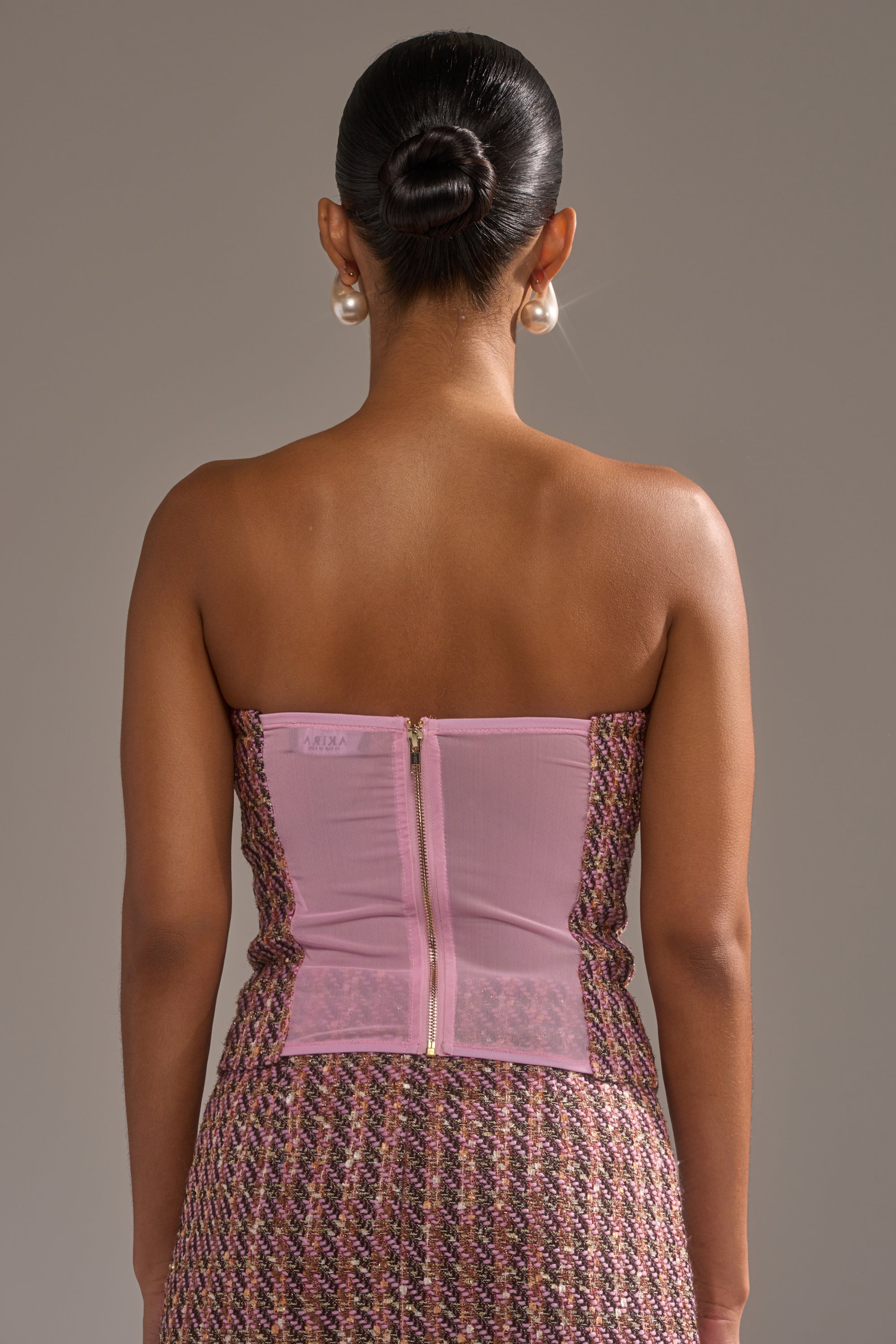 A woman with dark hair in a bun, wearing pearl earrings and the strapless TRUTH OR DARE TWEED CORSET dress with a visible back zipper, stands facing away from the camera against a plain background. 3