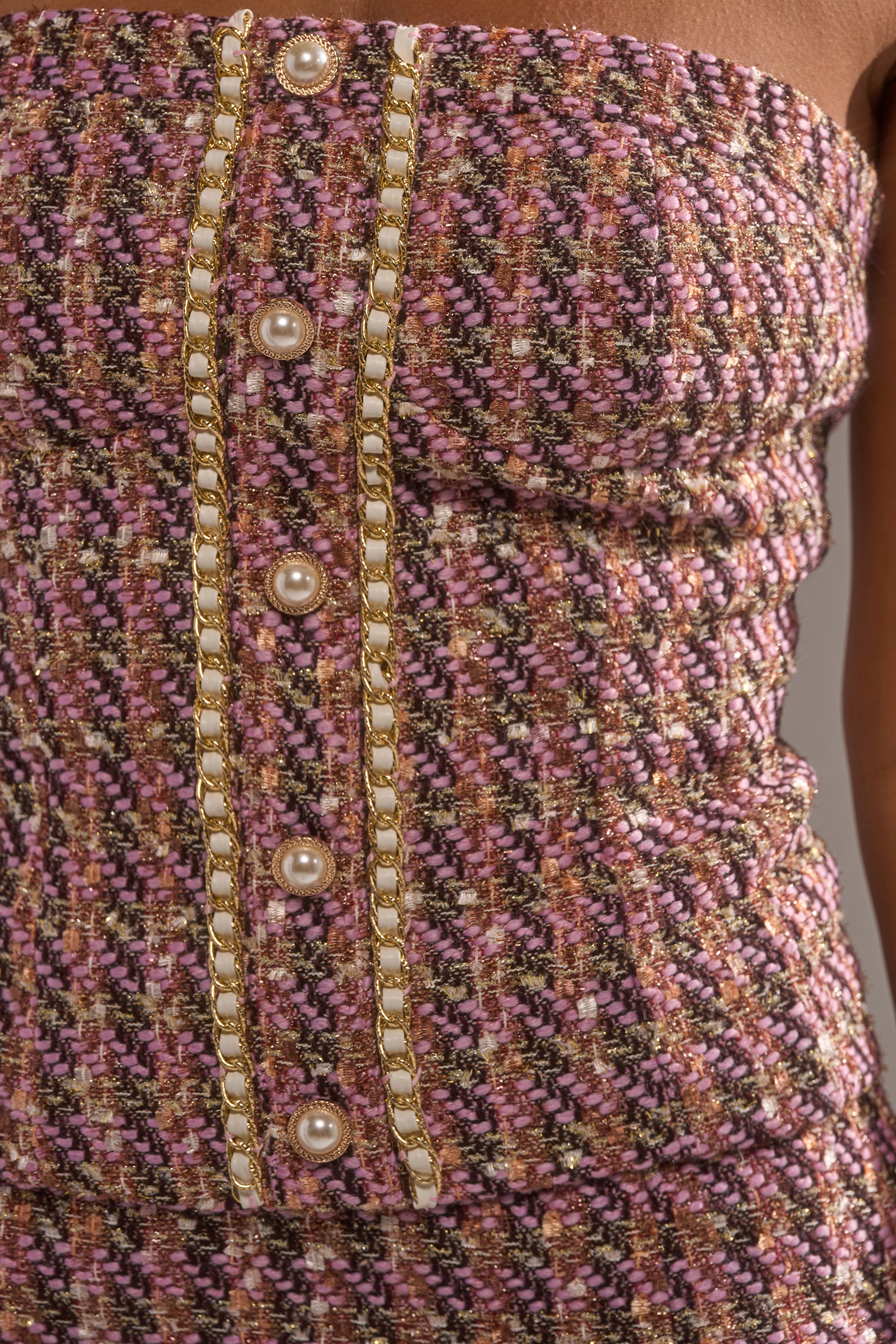 A close-up of a strapless, textured dress in pink, purple, and gold with vertical gold chains and pearl buttons, featuring a tweed corset top inspired by the TRUTH OR DARE TWEED CORSET. 6