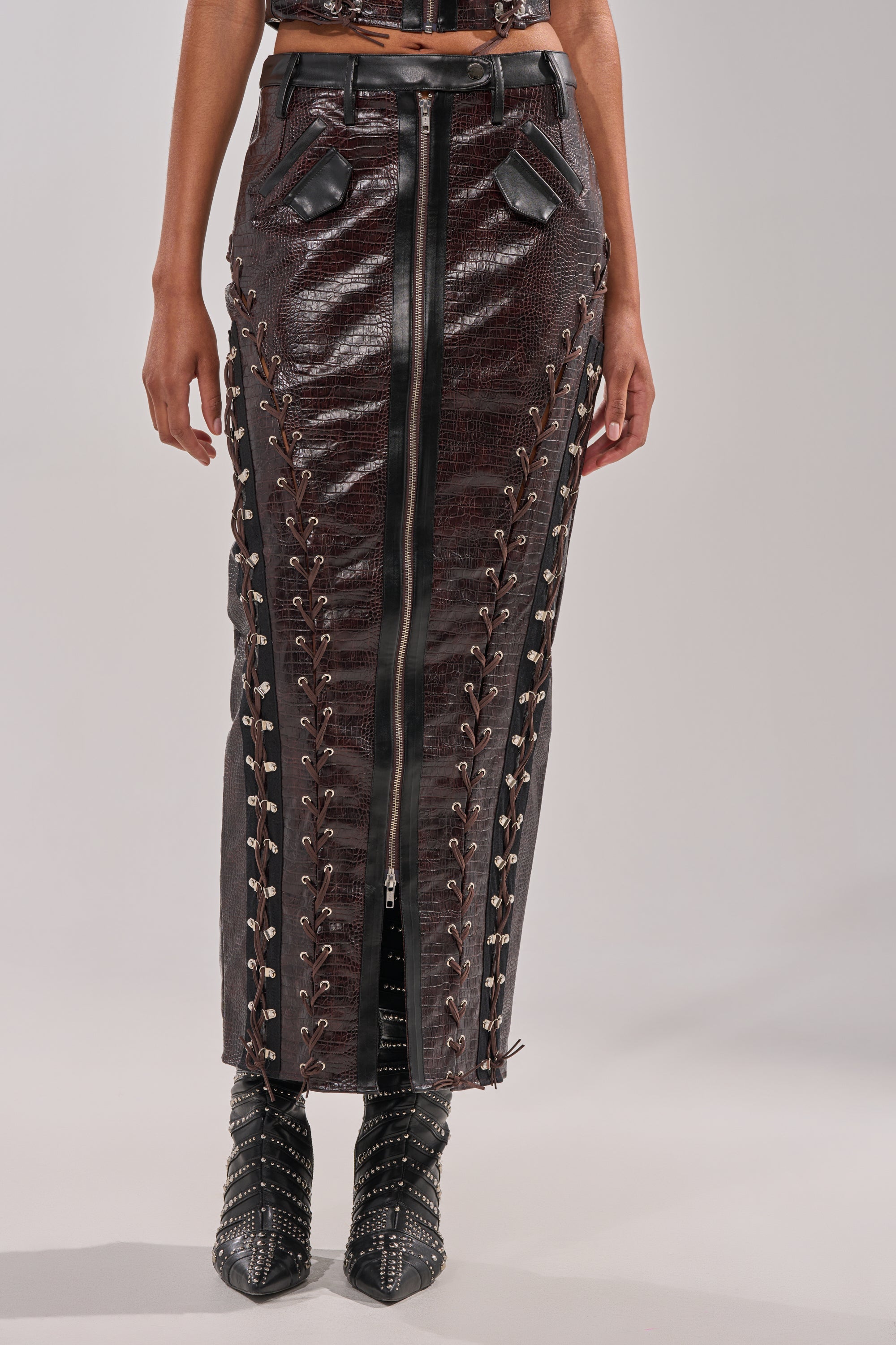 A person wears the TOO SEXY FOR YOU MAXI SKIRT, a high-waisted dark brown faux leather lace-up skirt with zipper front and faux pocket flaps, paired with black studded pointed boots. The background is plain light gray.