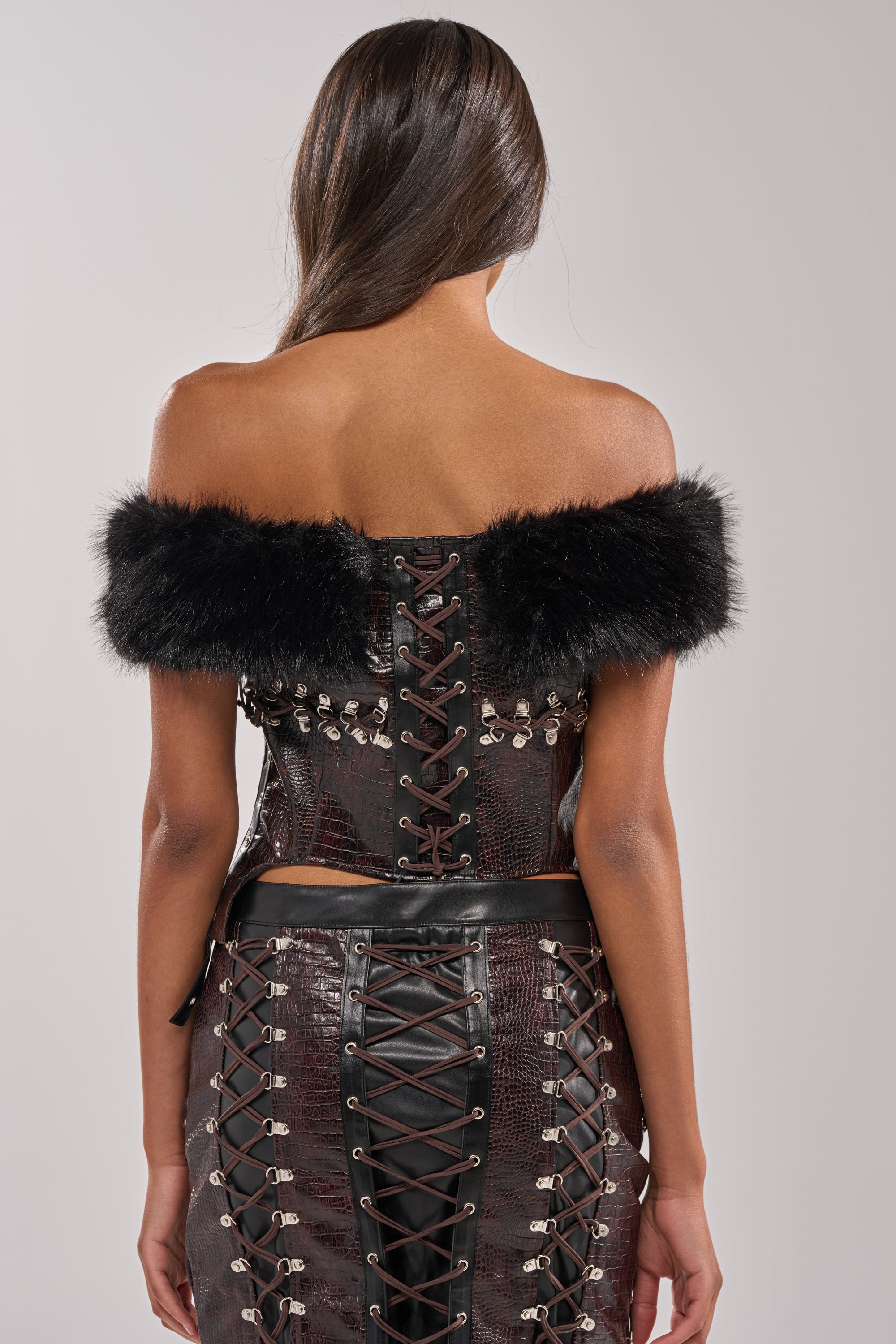 A woman with long brown hair faces away, wearing the TOO SEXY FOR YOU CORSET—an off-the-shoulder faux leather top with black faux fur trim and crisscross lace-up back—paired with a matching high-waisted skirt. 3