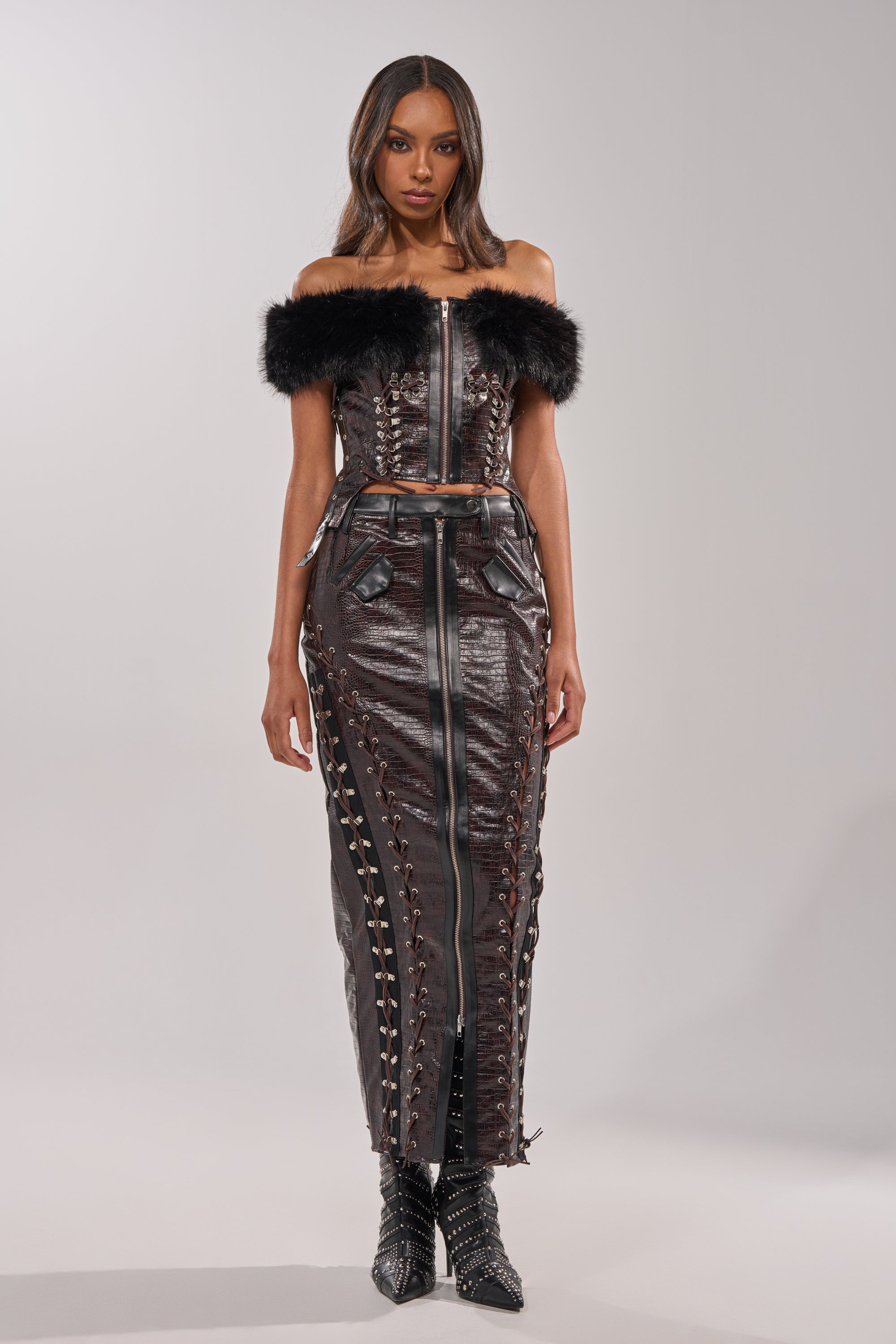 A woman faces forward in the TOO SEXY FOR YOU CORSET, a dark off-the-shoulder faux leather top with black faux fur trim, paired with a matching high-waisted lace-up skirt and pointed black boots. She has long, wavy hair. 4