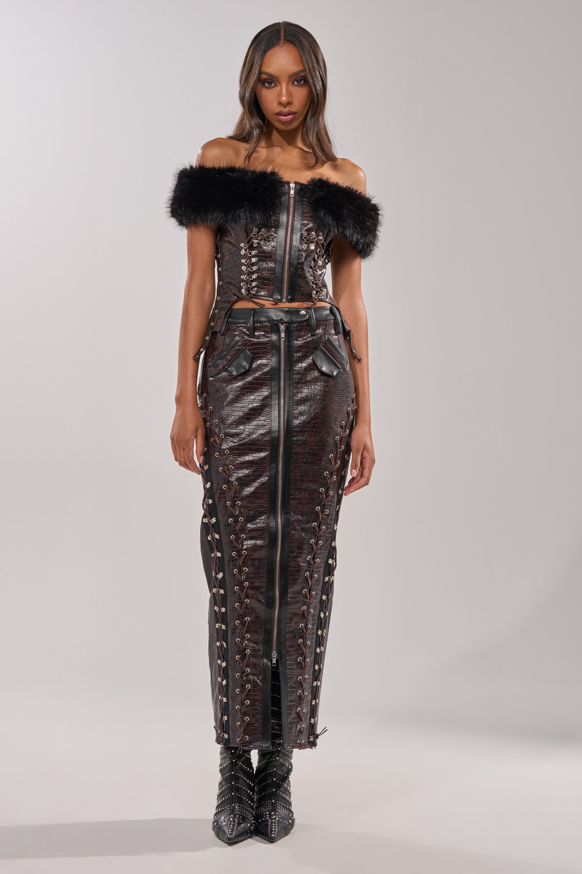 A woman faces forward in the TOO SEXY FOR YOU CORSET two-piece, featuring off-the-shoulder design, dark textured fabric, faux fur trim, and lace-up accents. She pairs it with pointed black boots against a simple, light background. 5