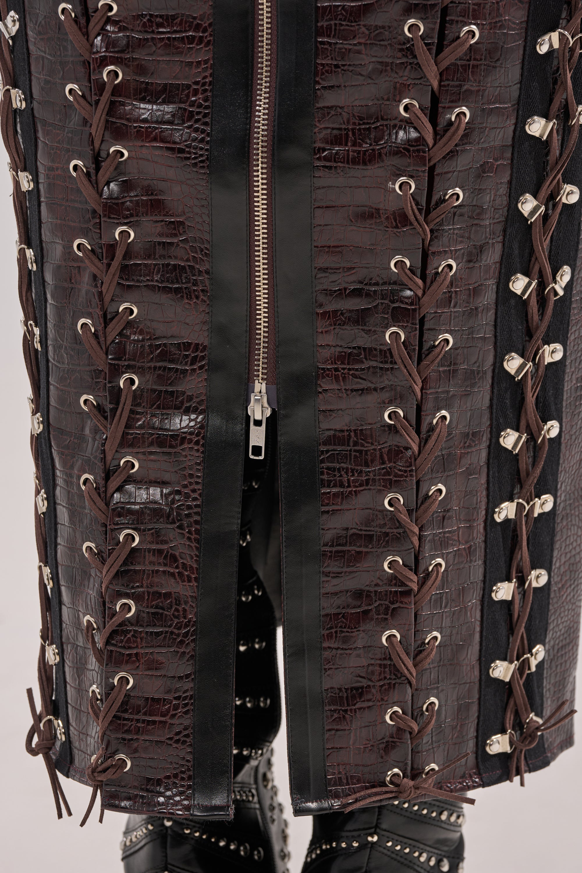 Close-up of the TOO SEXY FOR YOU CORSET in dark brown, crocodile-patterned faux leather with black strips, silver eyelets, crisscrossed lacing, center zipper, and studded boots peeking out at the bottom. 8