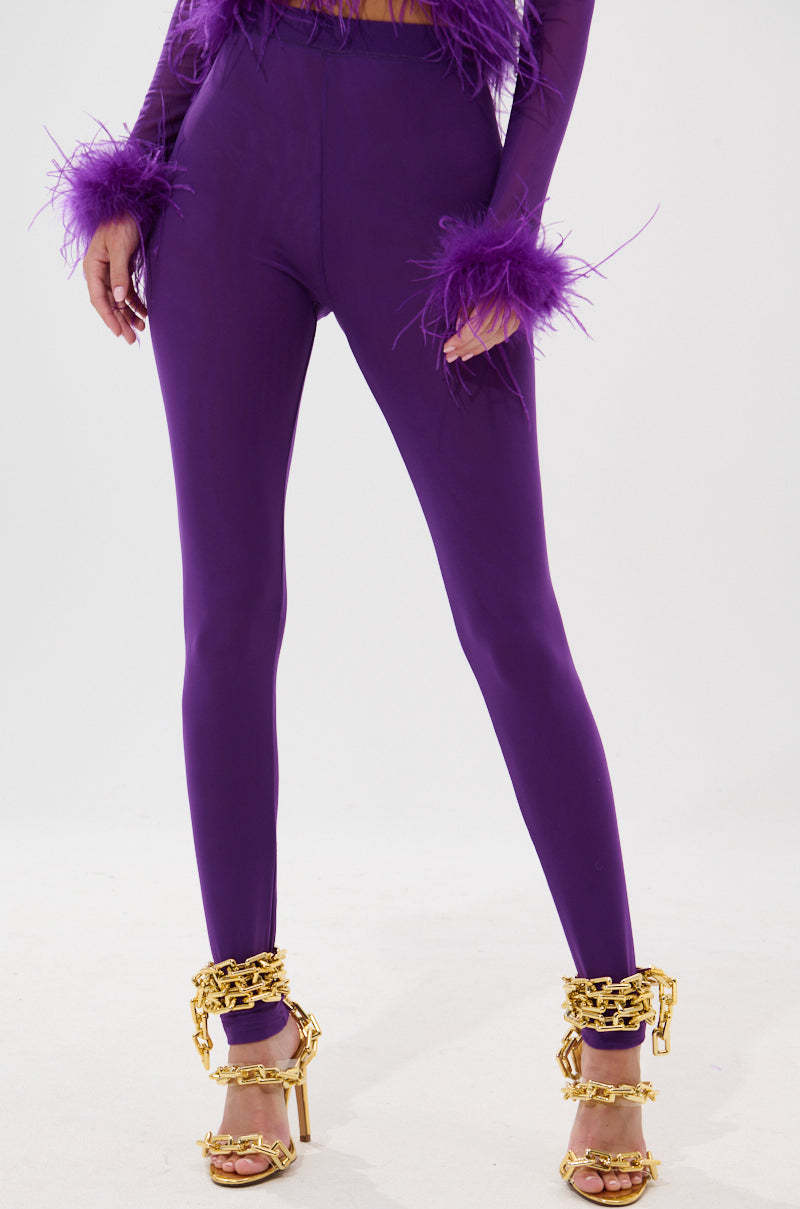 A person wears the PUMPED UP MESH LEGGING in purple with a matching feather-cuff top and gold heels with chunky chain details, standing against a plain white background. 1