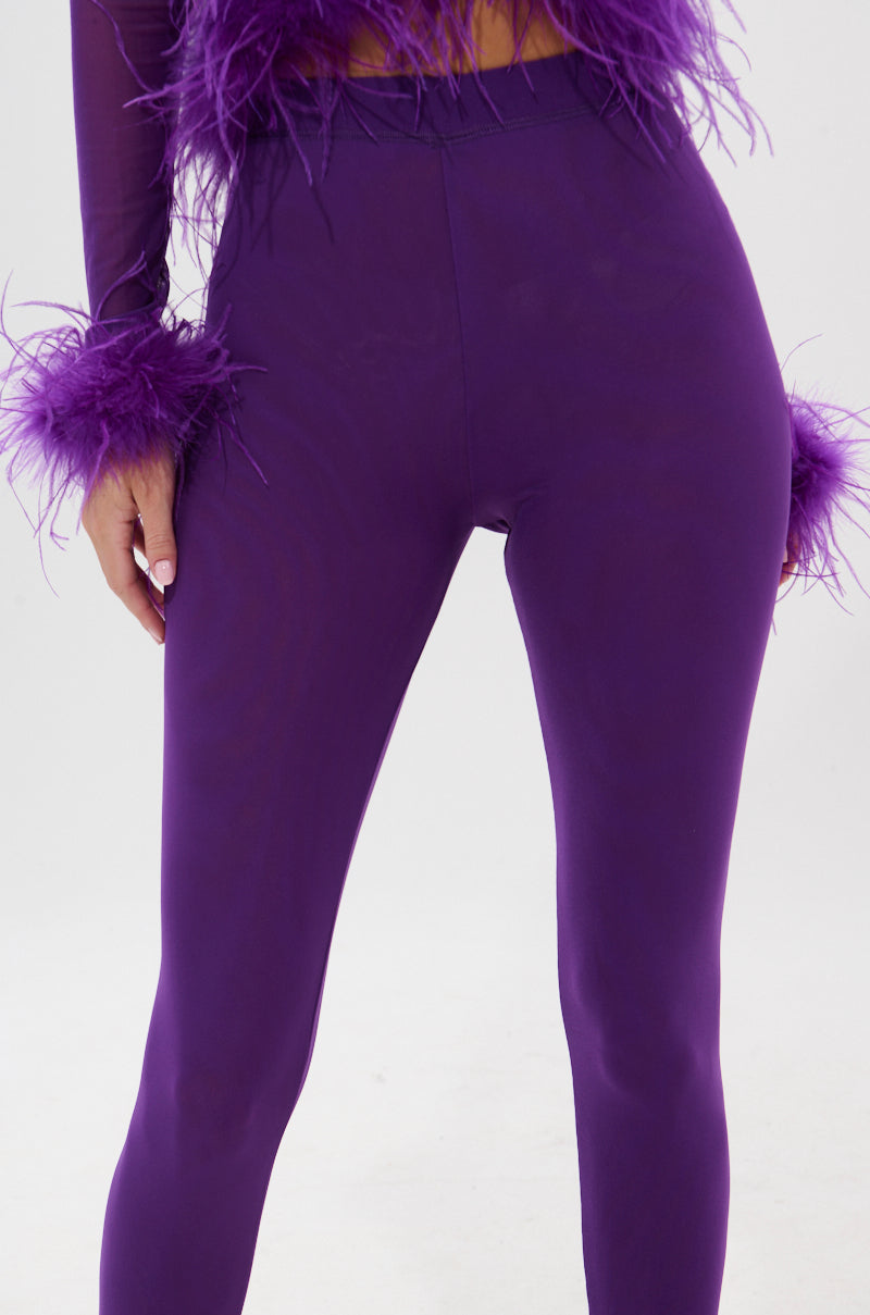 A person wears the PUMPED UP MESH LEGGING in high-rise royal purple with feather trim at the wrists and hip, standing against a plain white background. 7