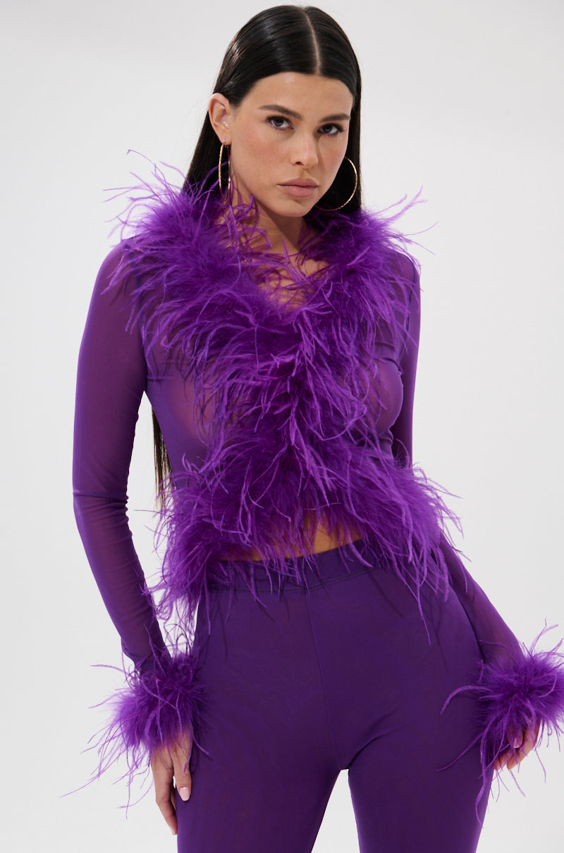 A woman wears the PUMPED UP FEATHER BLOUSE, a fitted purple top with long sleeves and feather details, paired with matching pants. She accessorizes with hoop earrings and has straight dark hair and a confident look.