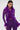 A woman wears the PUMPED UP FEATHER BLOUSE, a fitted purple top with long sleeves and feather details, paired with matching pants. She accessorizes with hoop earrings and has straight dark hair and a confident look.
