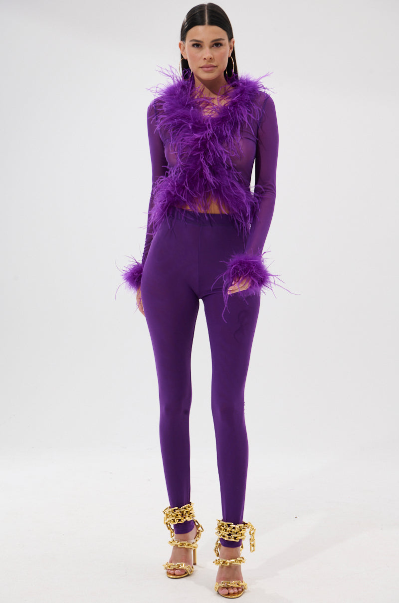 A woman poses against a white background in the PUMPED UP FEATHER BLOUSE, a vibrant purple top with feather trim on the sleeves, paired with gold high-heeled sandals adorned with bold chain accents.