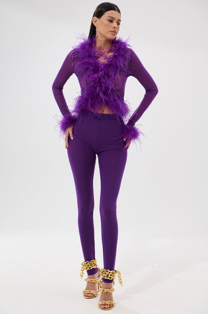 A woman poses confidently in a fitted, bright purple PUMPED UP FEATHER BLOUSE with feather trim on the top and wrists, paired with gold chain high-heeled sandals against a plain white background.