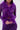 A woman wearing the PUMPED UP FEATHER BLOUSE, a sheer mesh top with fluffy purple feather trim on the neckline and cuffs, pairs it with matching pants and holds her hands together in front of her.