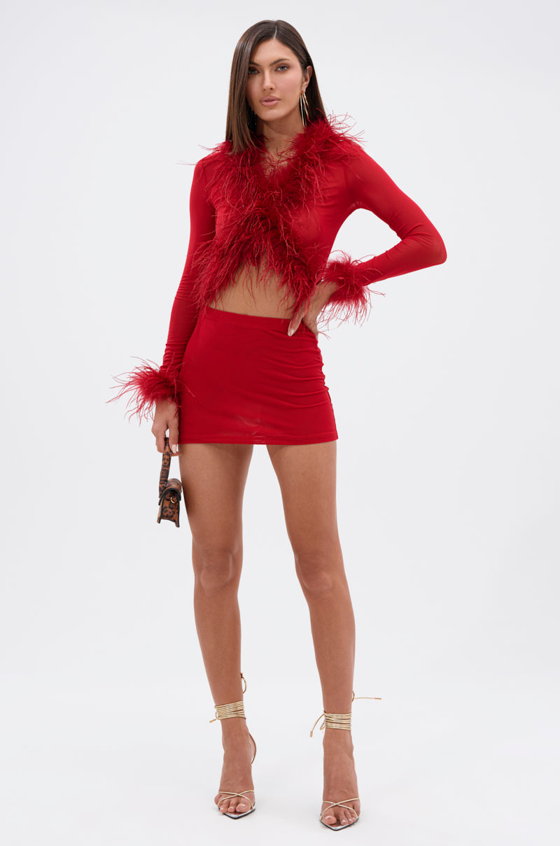 A woman wears the FIERCE MESH MINI SKIRT, a curve-hugging red high-rise mini, paired with a matching long-sleeve crop top featuring feather trim. She holds a clutch and sports gold strappy heels against a plain white background.