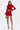 A woman wears the FIERCE MESH MINI SKIRT, a curve-hugging red high-rise mini, paired with a matching long-sleeve crop top featuring feather trim. She holds a clutch and sports gold strappy heels against a plain white background.